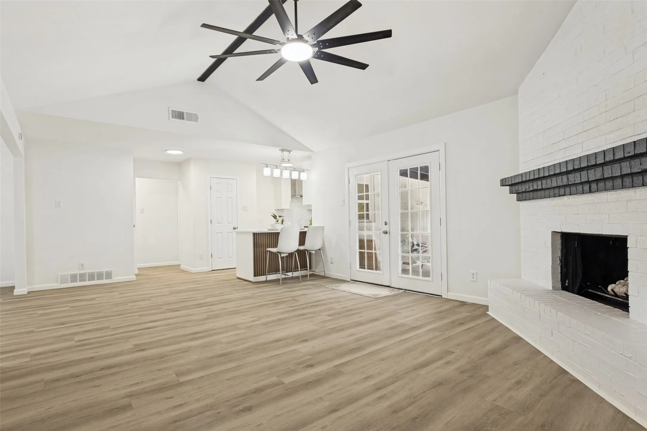 Vaulted ceiling with a contemporary ceiling fan, painted brick fireplace with a dark mantel, wood-finish flooring, and double French doors with grid-patterned glass