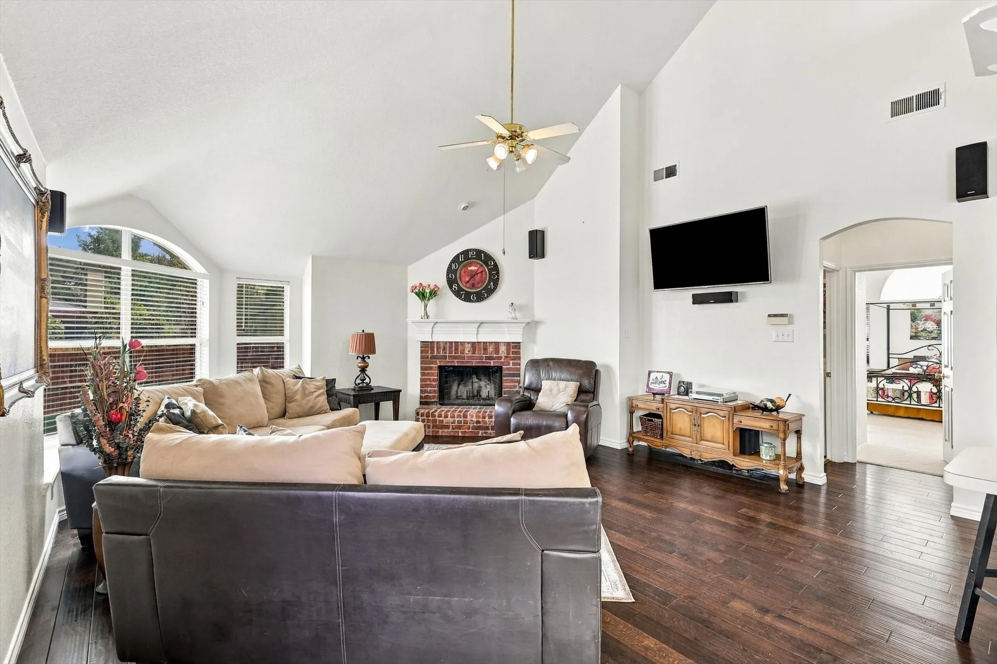 Vaulted ceilings, wood-finish flooring, brick-surround fireplace, arched window, and ceiling fan