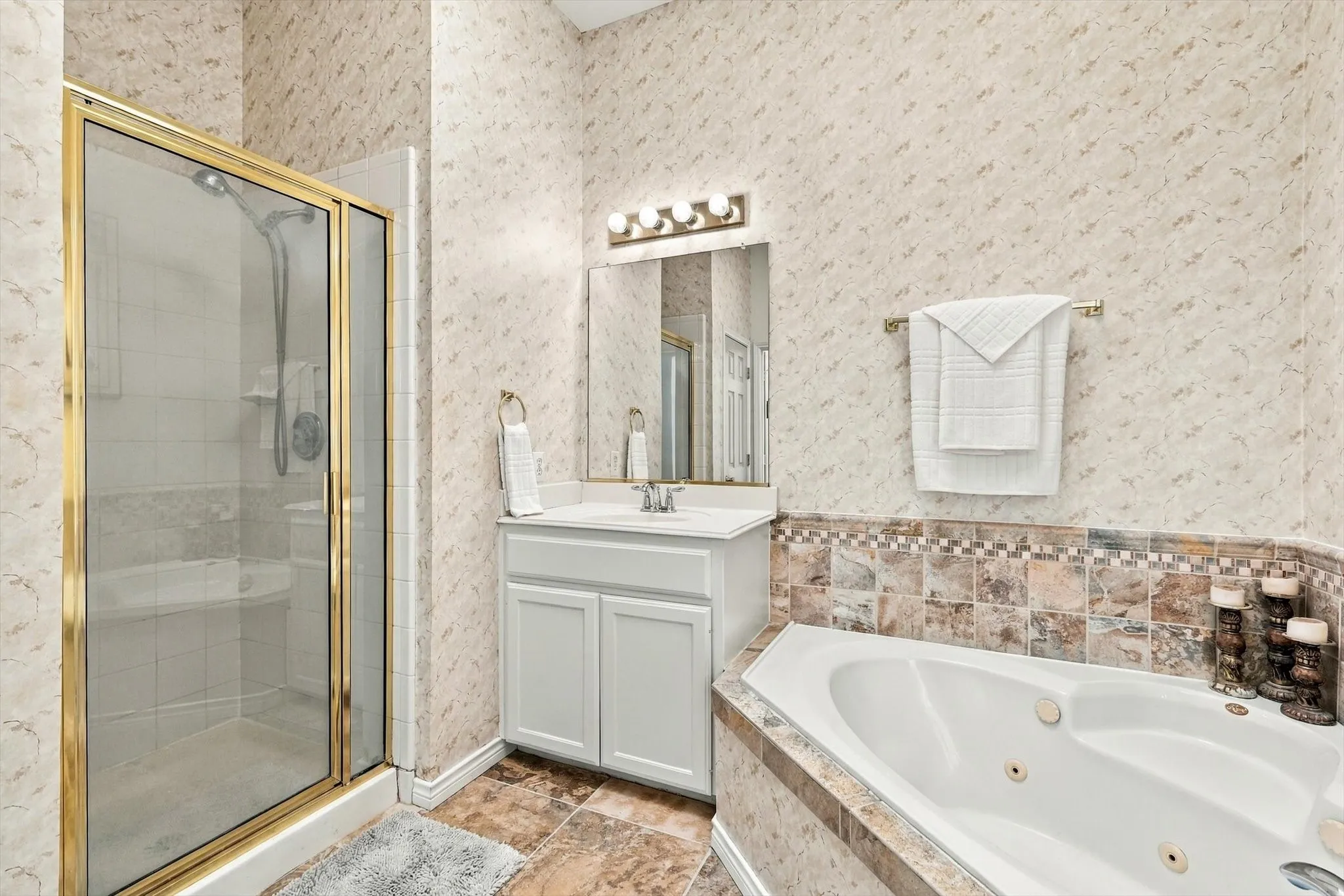 Shower with gold-toned frame and clear glass enclosure, single vanity with white cabinetry, built-in mirror with integrated lighting, tiled flooring, and corner jetted bathtub with tiled surround