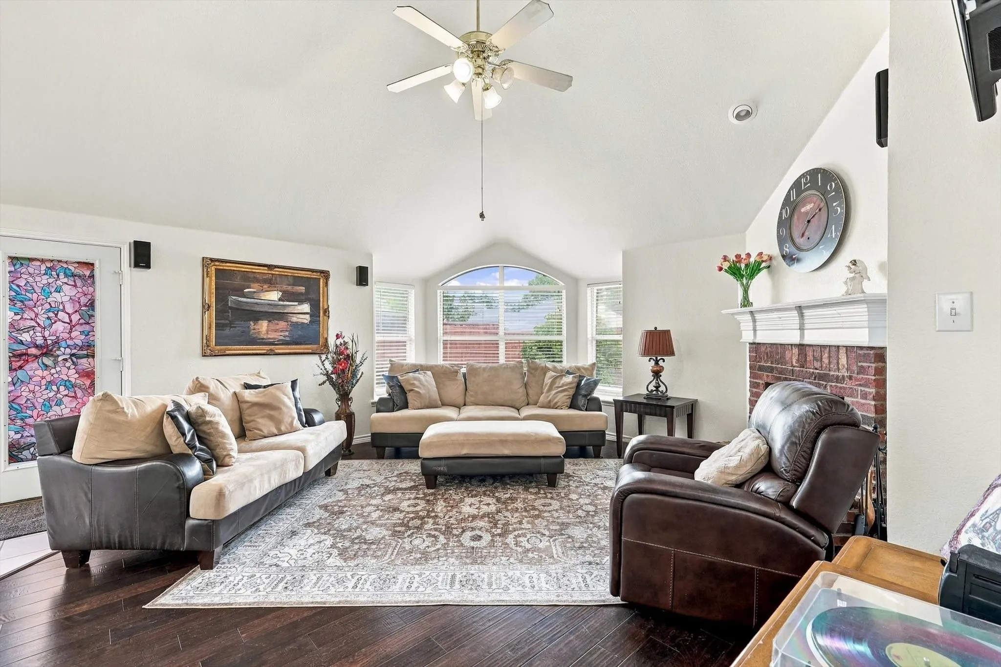 Vaulted ceiling with a ceiling fan, dark wood-finish flooring, brick fireplace with a white mantel, arched window, and recessed lighting