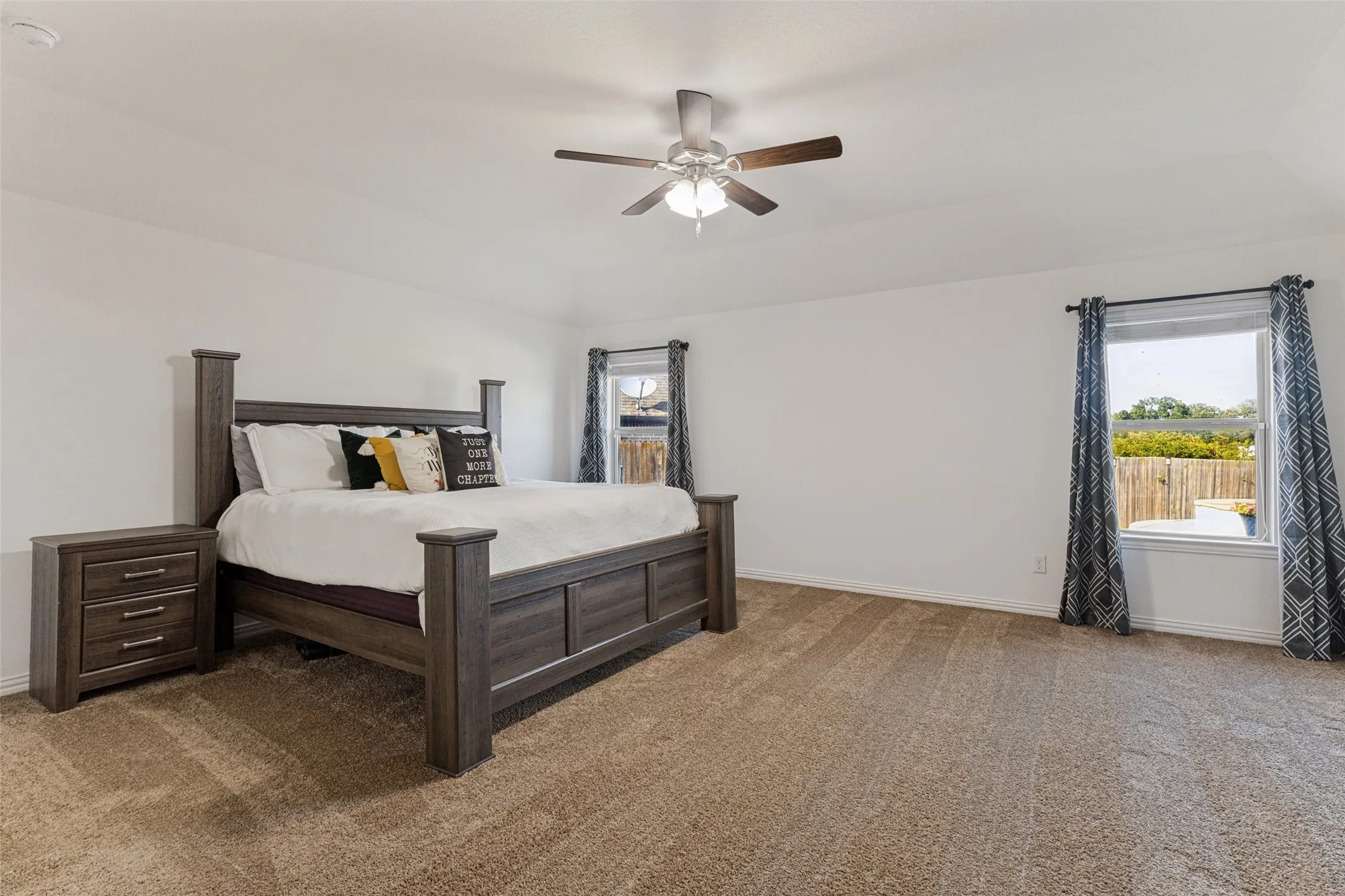 Spacious room featuring carpet flooring, two windows, a ceiling fan with integrated lighting, and white walls