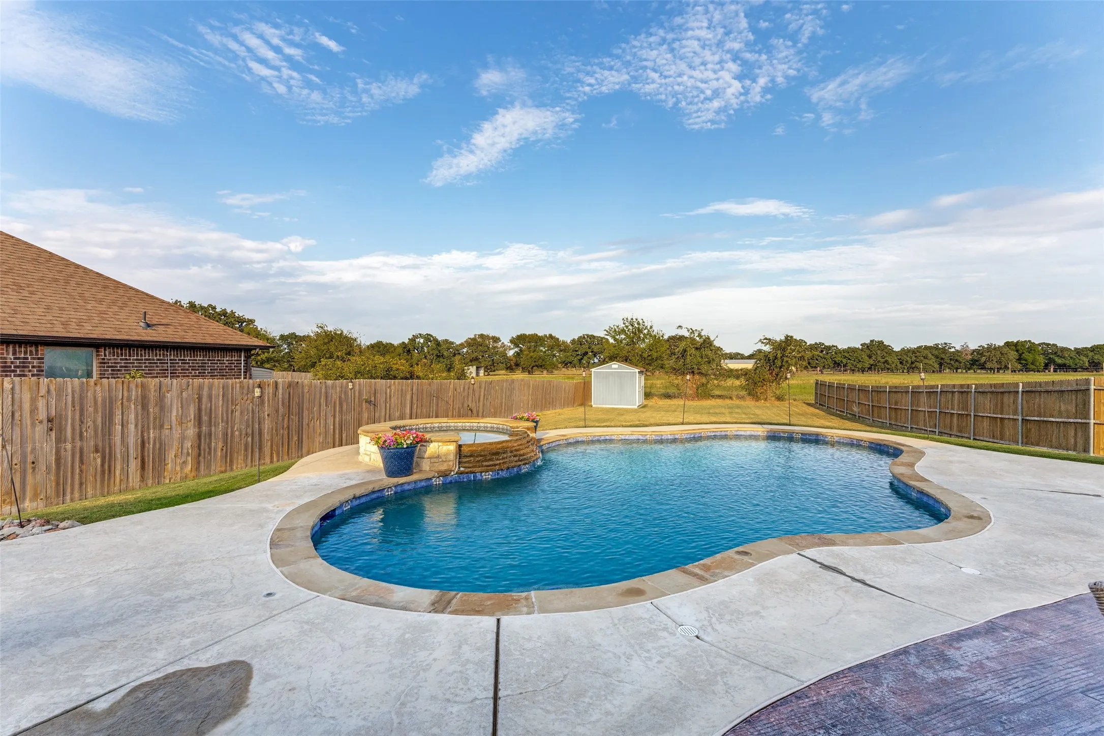Curved swimming pool with an integrated spa, expansive concrete decking, and perimeter fencing