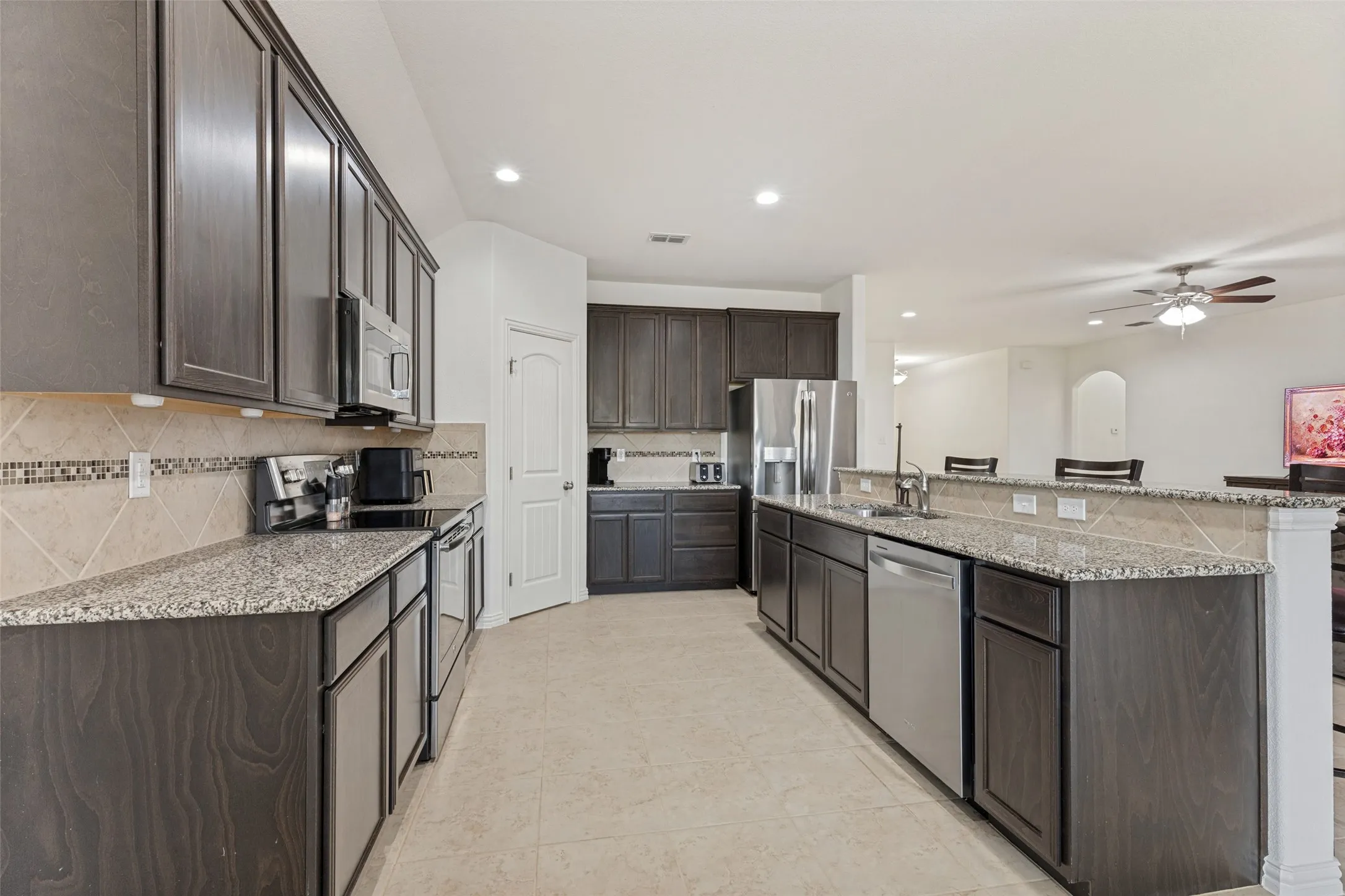 Kitchen featuring dark wood-finish cabinetry, light speckled countertops, stainless steel appliances, a tile backsplash, and recessed lighting
