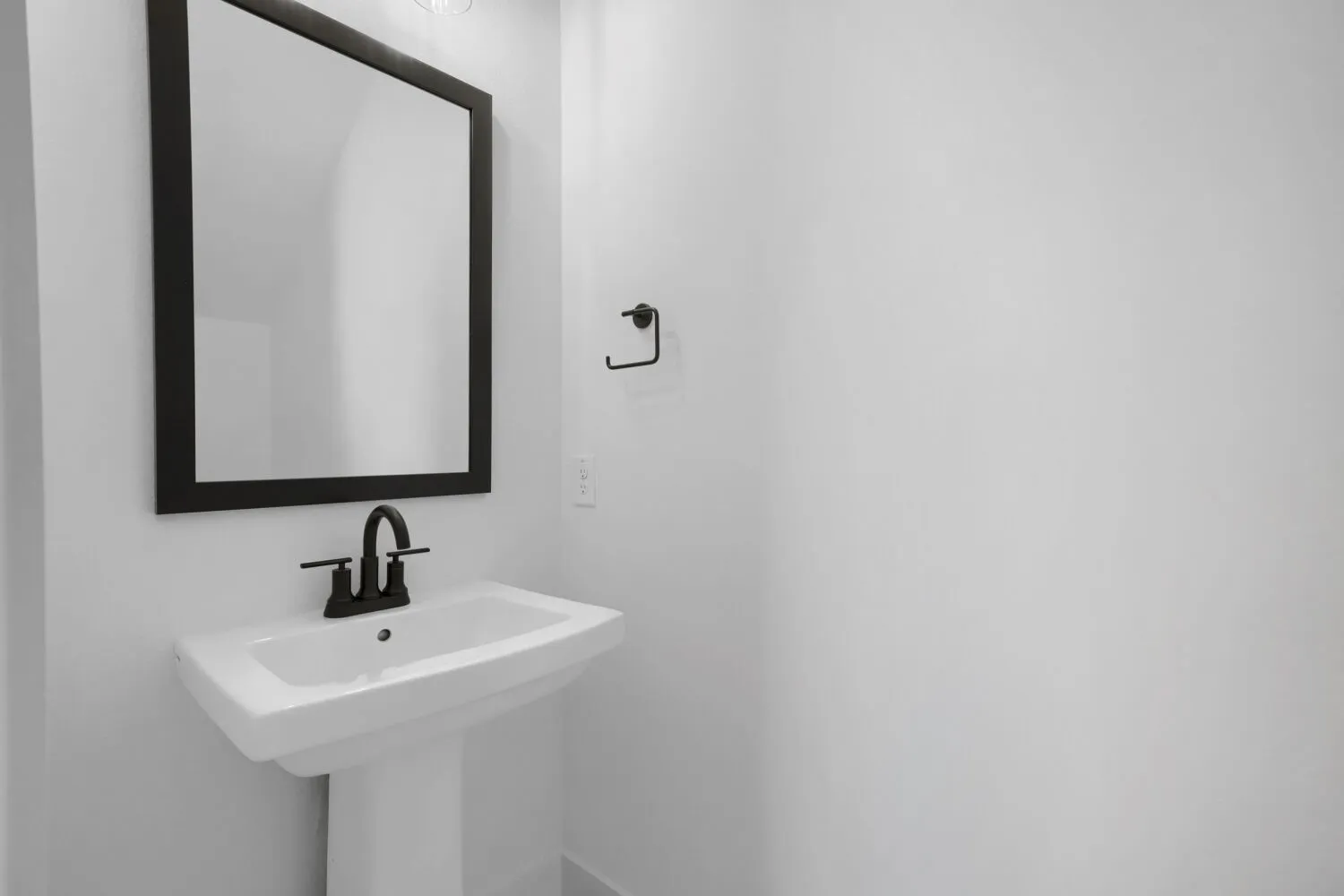 White pedestal sink with a matte black faucet and a framed wall mirror