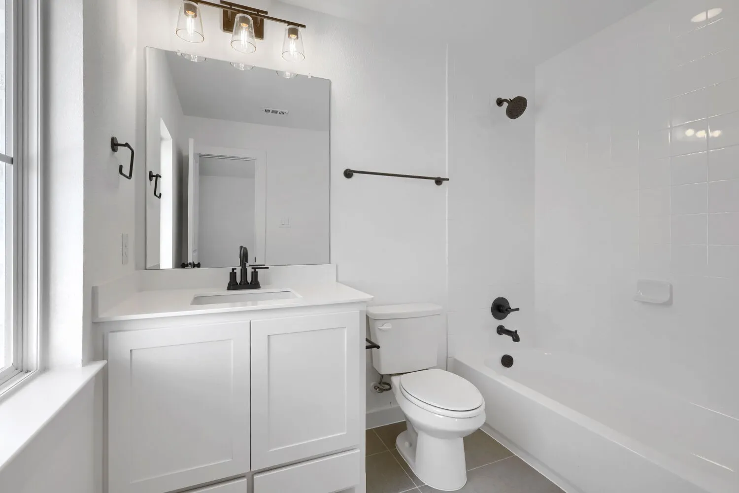 Modern bathroom featuring a white vanity with dual-sink configuration, large framed mirror with integrated lighting, ceramic tile surround, and a combined shower-tub with glass enclosure and matte black hardware