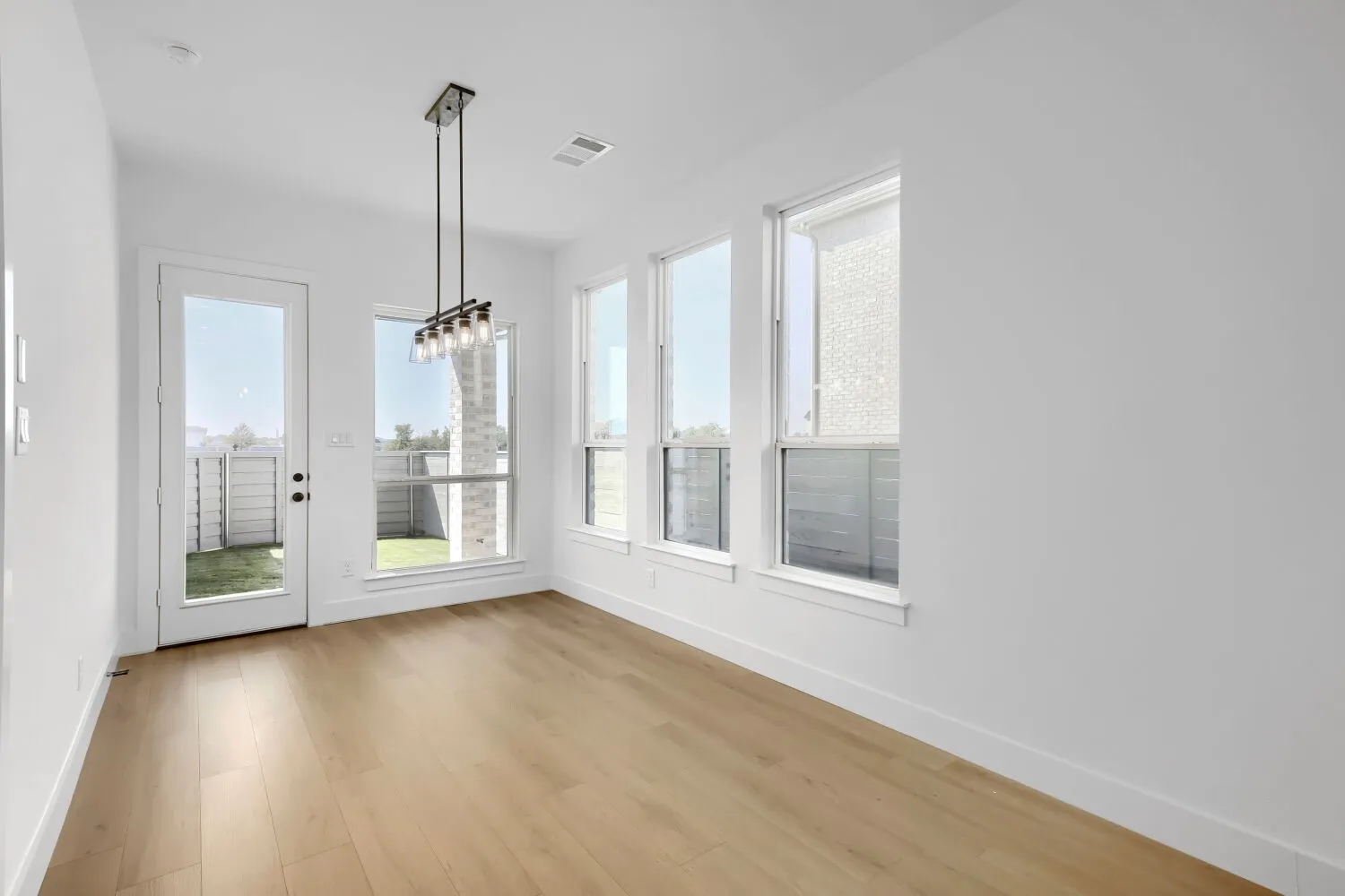 Bright, light-filled room showcasing expansive windows with contemporary framing, warm wood-finish flooring, crisp white walls, and a modern pendant light fixture suspended from the ceiling