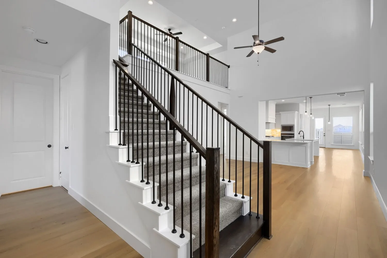 Open-concept living area featuring a dramatic staircase with black metal railings and white balusters, wide-plank wood-finish flooring throughout, recessed ceiling lighting, and a ceiling fan