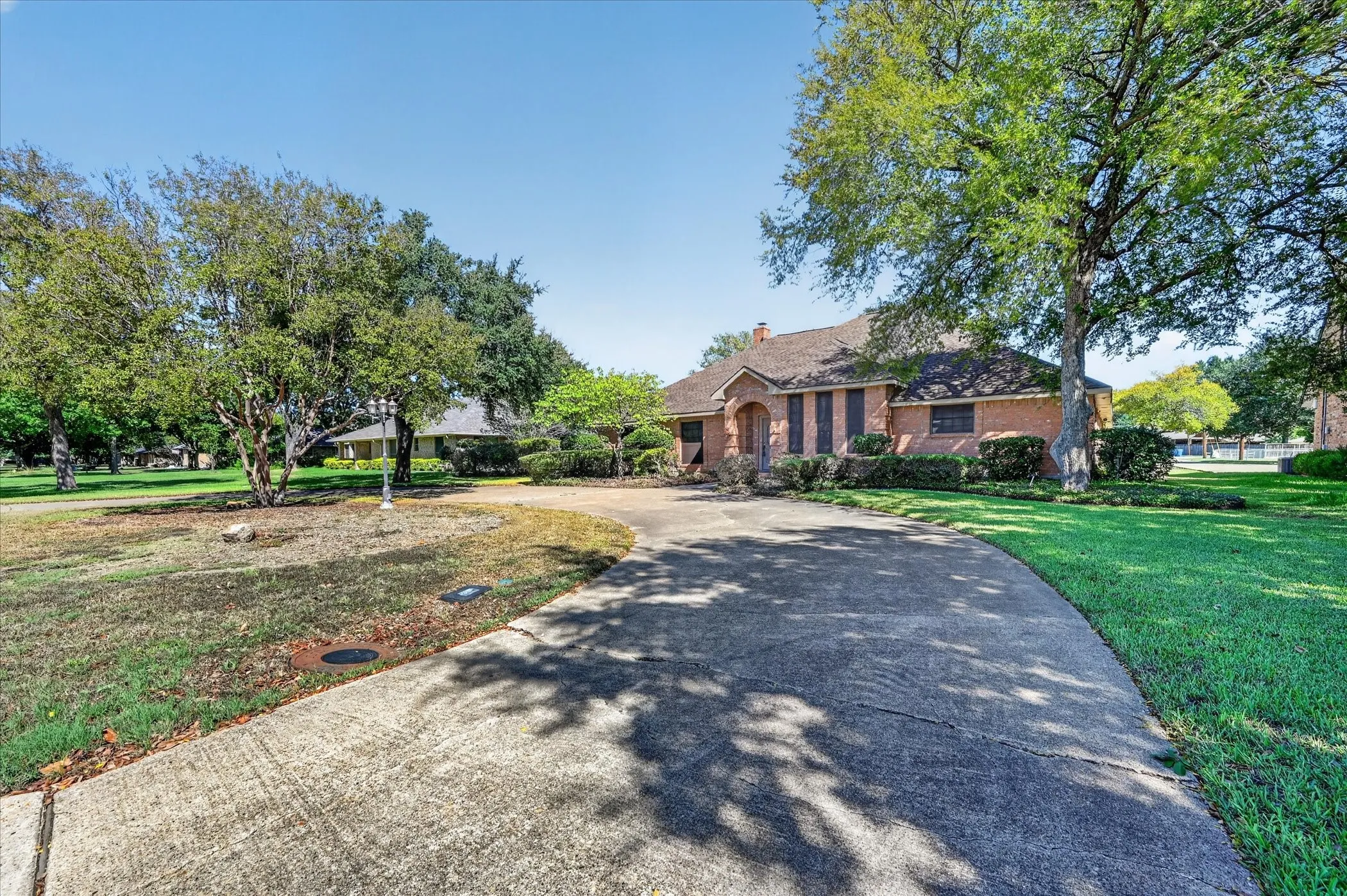 Curved concrete driveway with mature trees, brick exterior, and landscaped foundation plantings