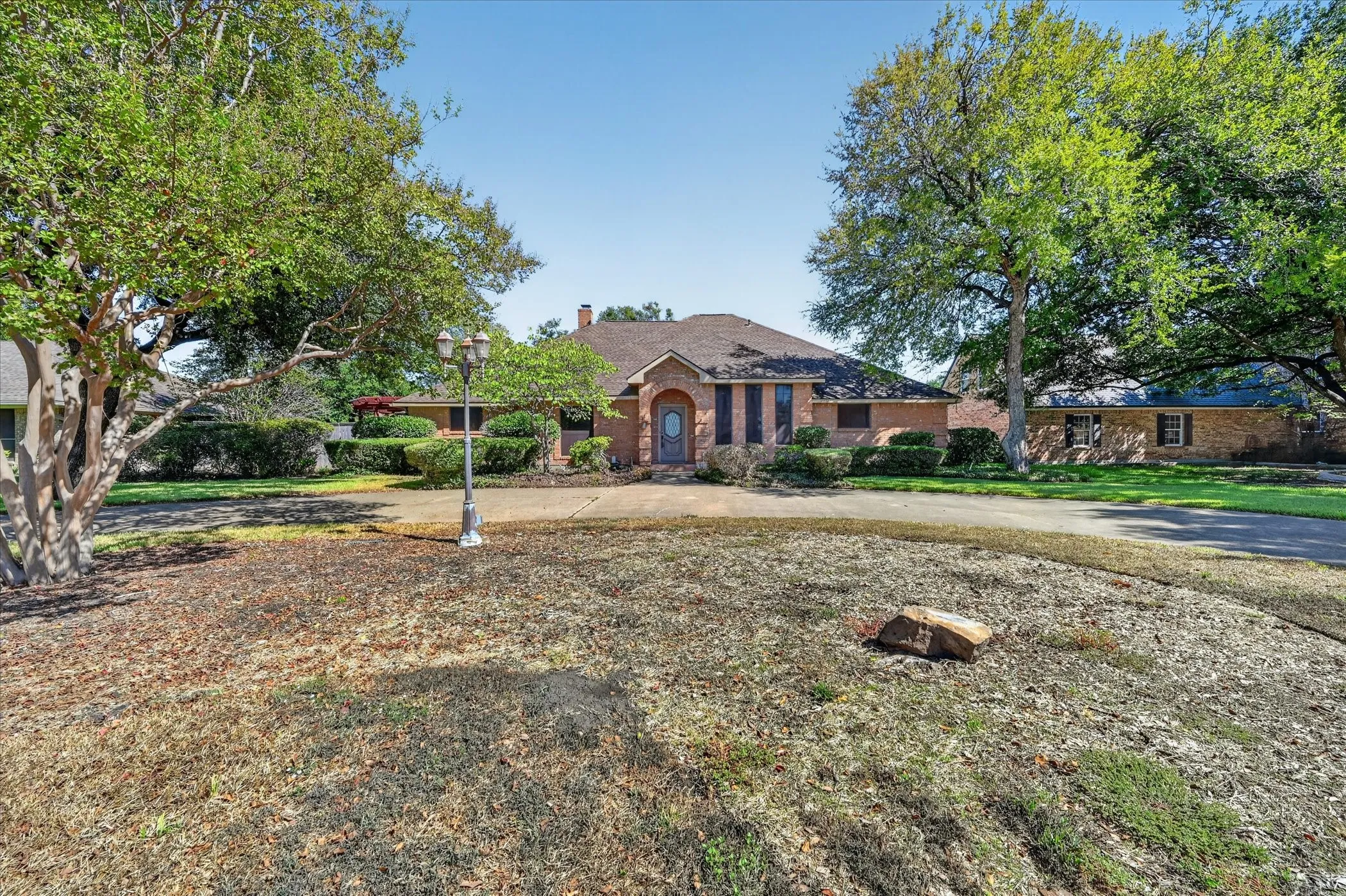 Brick exterior, arched entryway, dark-colored front door, decorative shutters, and mature trees