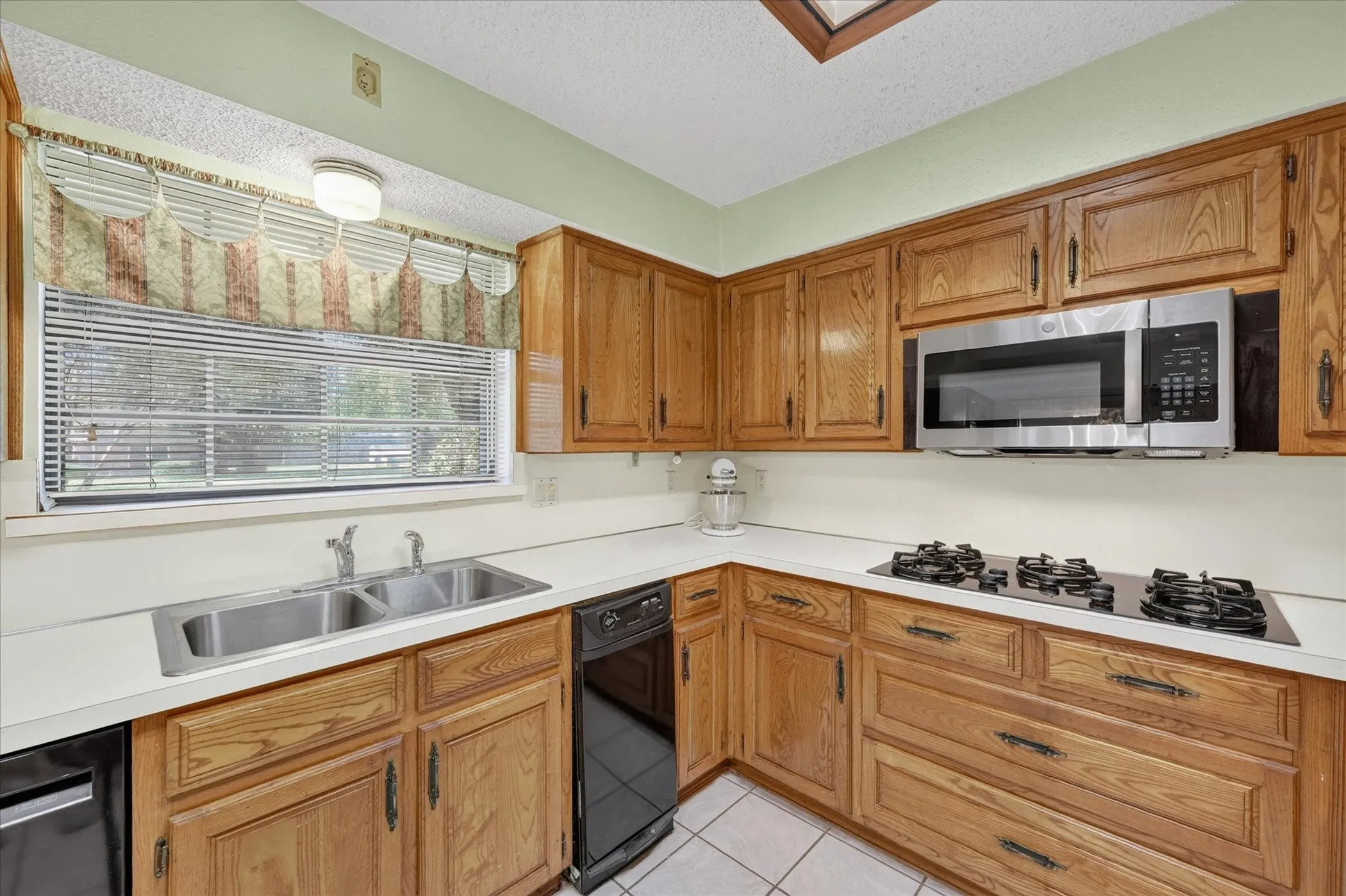 Kitchen featuring wood cabinetry, white countertops, a double basin stainless steel sink, a stainless steel microwave, and a four-burner cooktop