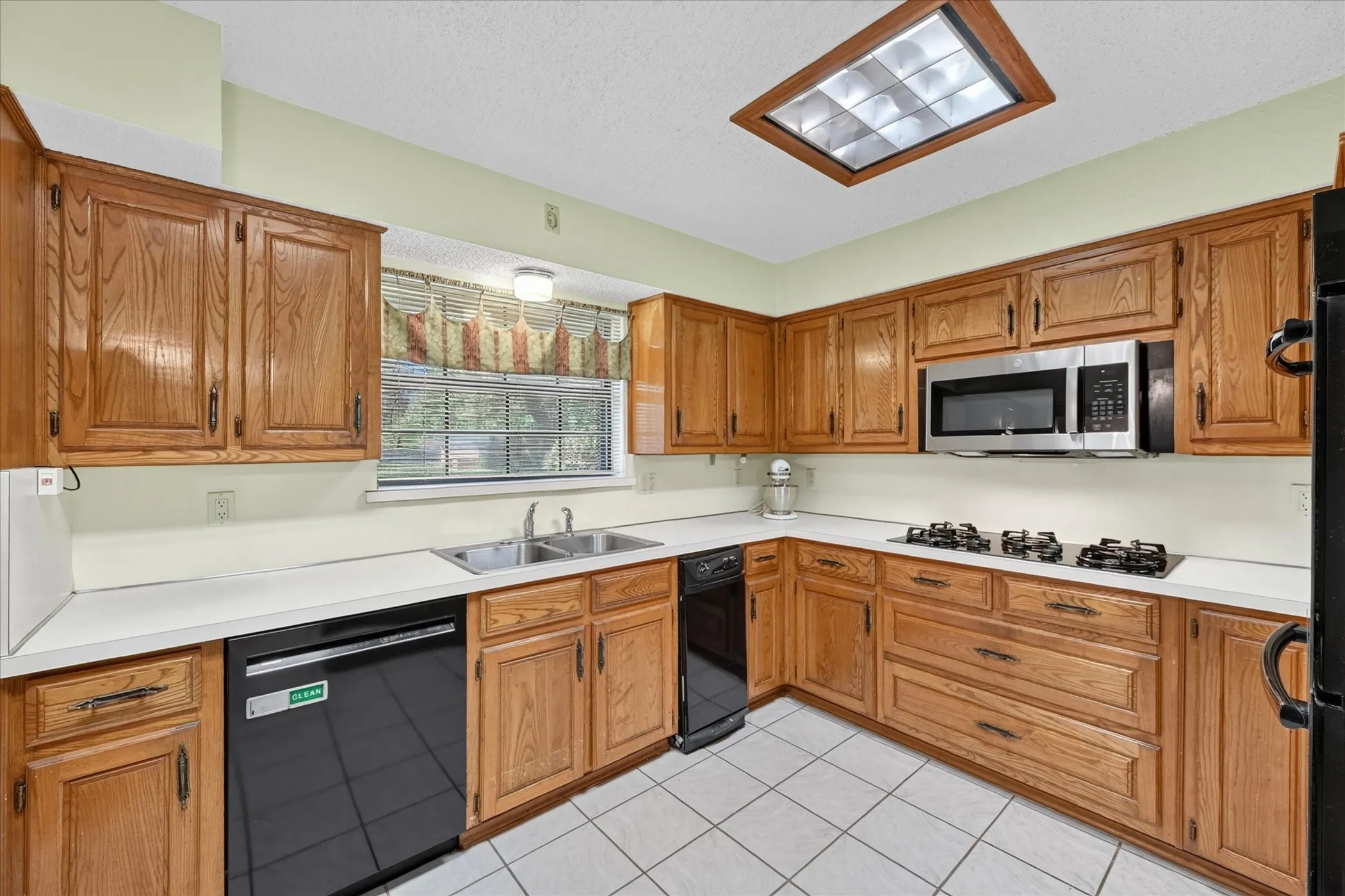 Kitchen featuring wood cabinetry, white countertops, a stainless steel microwave, black dishwasher, and white tile flooring