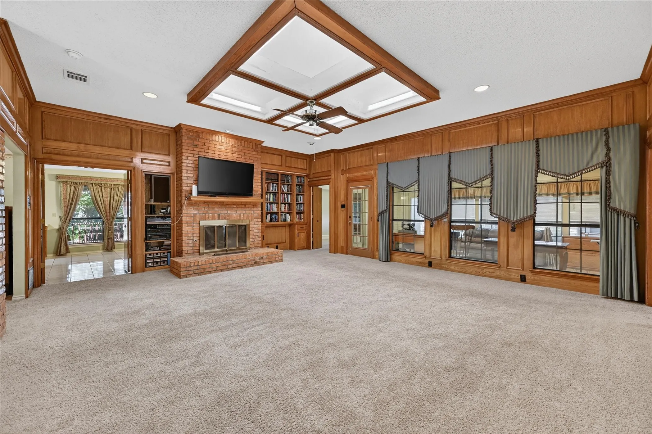 Carpeted living area featuring a brick fireplace, built-in shelving, wood wall paneling, and a coffered ceiling with skylights