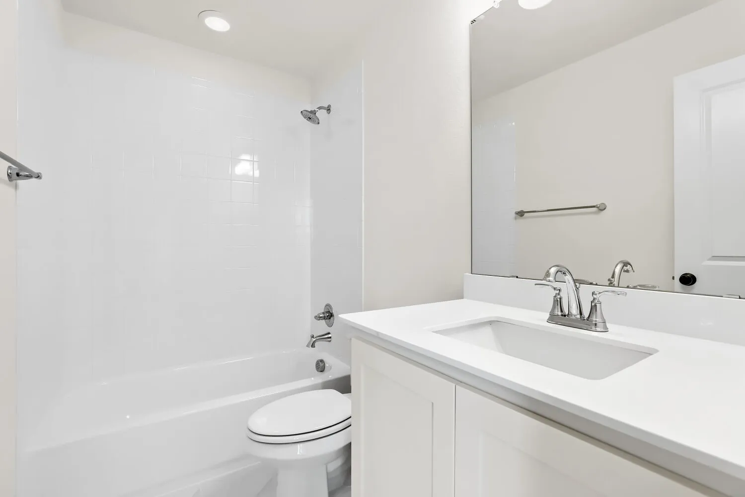 White vanity with an integrated sink and chrome faucet, large frameless mirror, and a white bathtub with a tiled shower surround