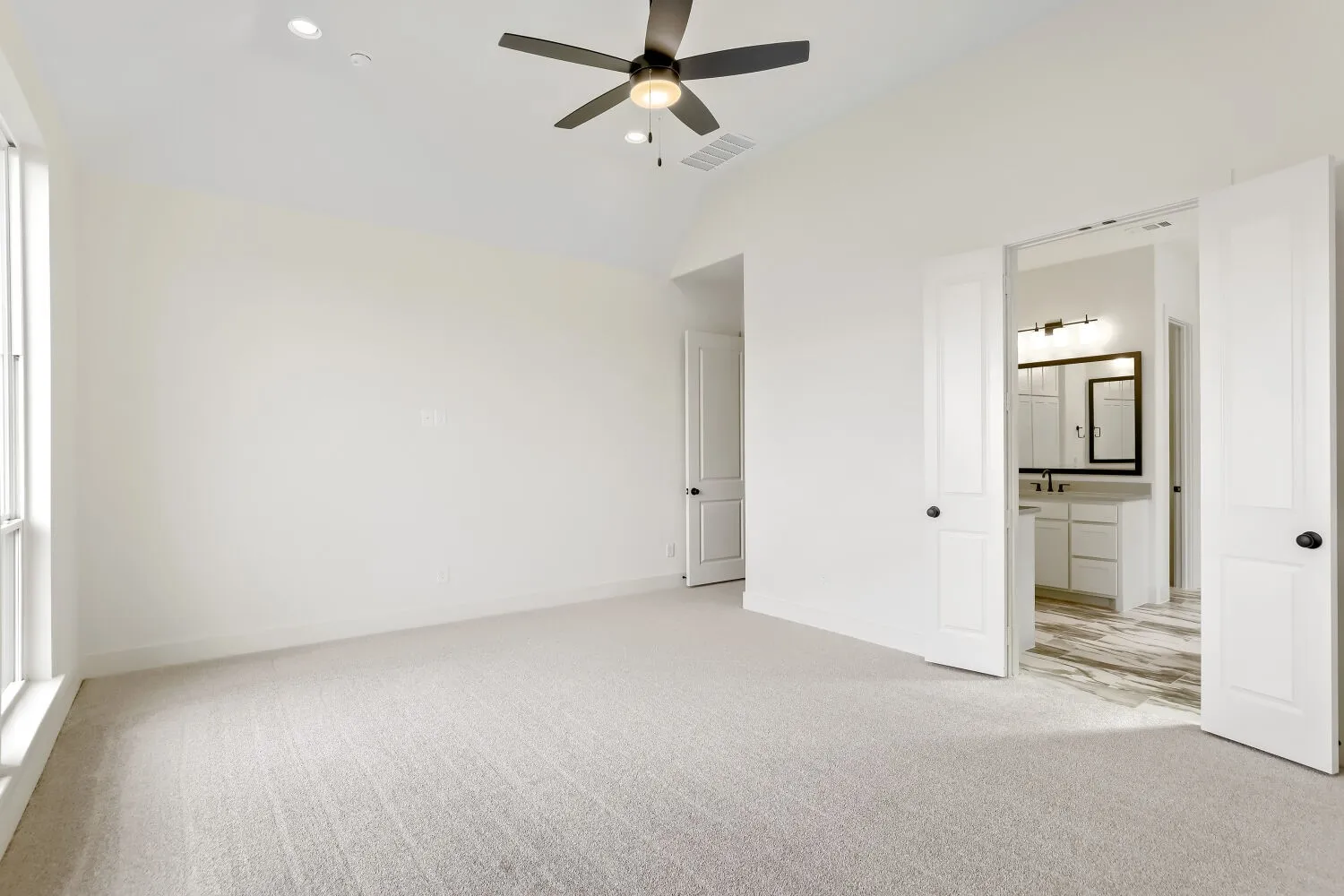 Vaulted ceiling with a ceiling fan, recessed lighting, carpet flooring, a large window, and double doors opening to a bathroom with a vanity and wood-finish flooring