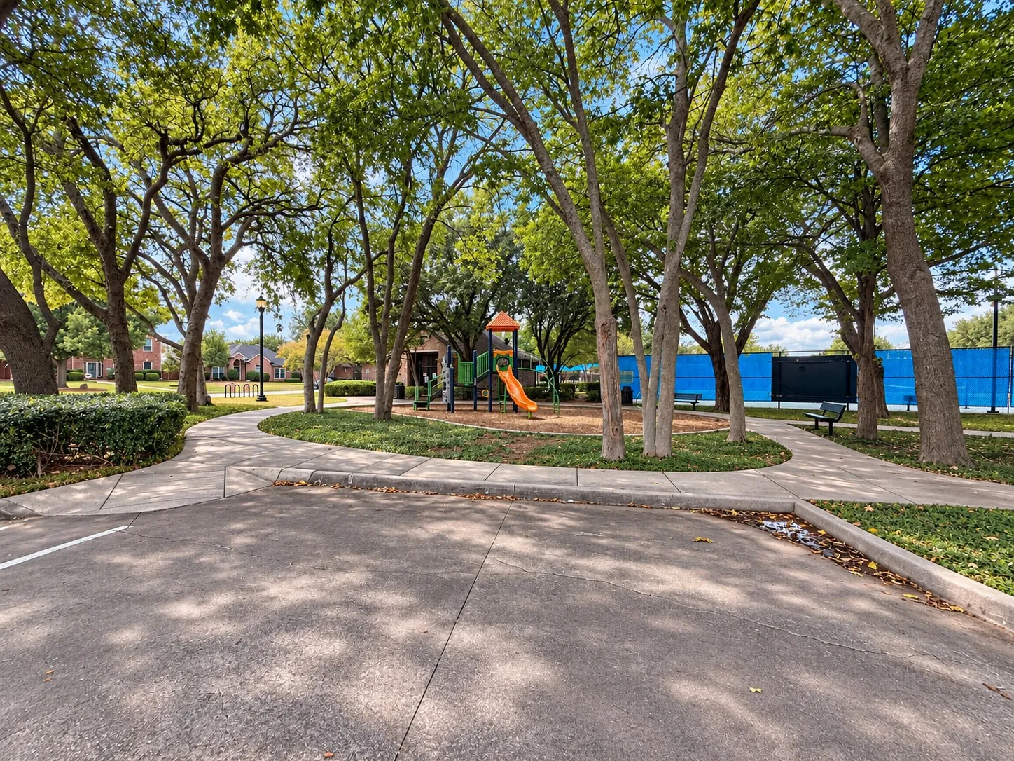 Paved walkways surrounded by mature trees and manicured landscaping, featuring a playground and a tennis court with blue fencing