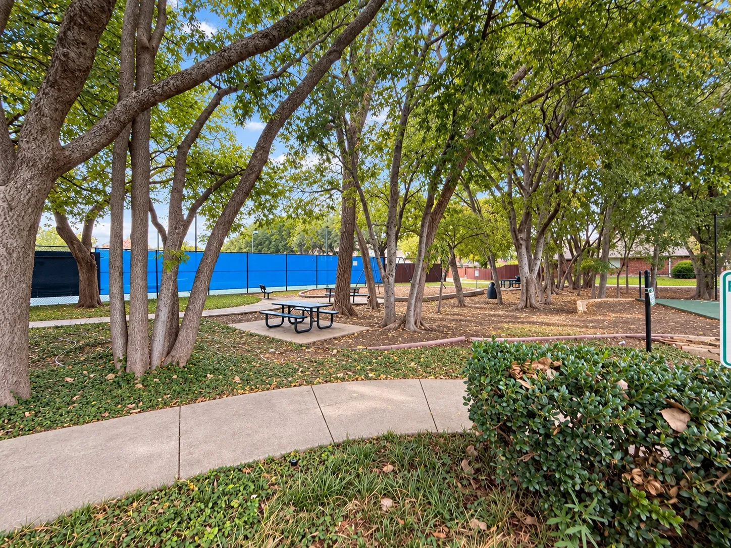 Tennis court with blue fencing, paved walking path, picnic tables, mature trees, and landscaped grounds
