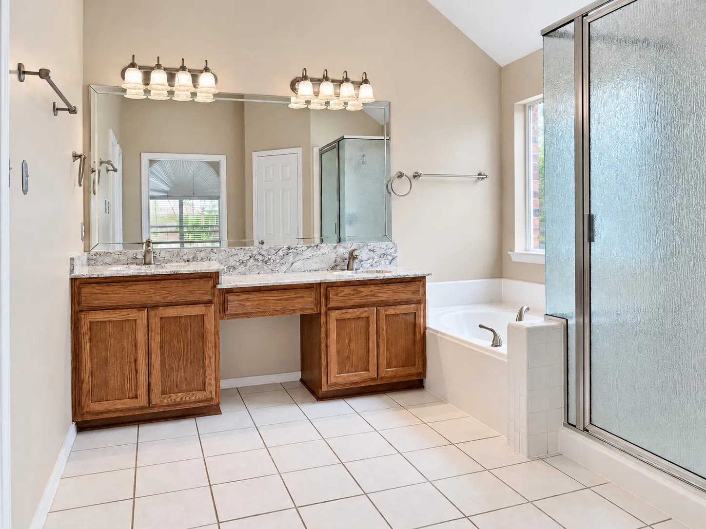 Dual wood-finish vanities with light stone countertops, integrated soaking tub, and a glass-enclosed shower