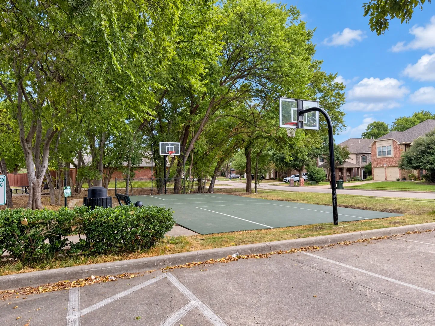 Recreational court featuring two basketball hoops, surrounded by mature trees and landscaping