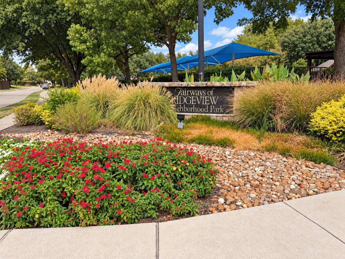 Stone-clad neighborhood park sign, mature trees, vibrant red flowering shrubs, and ornamental grasses