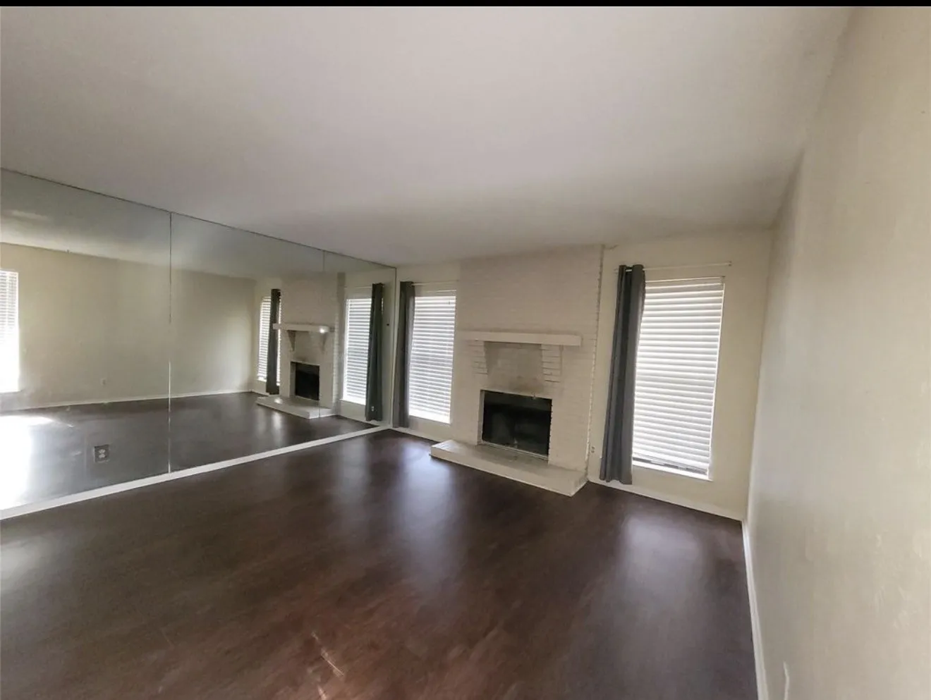 Wood-finish flooring, full-wall mirrored accent, brick fireplace with a mantel, and multiple windows with blinds