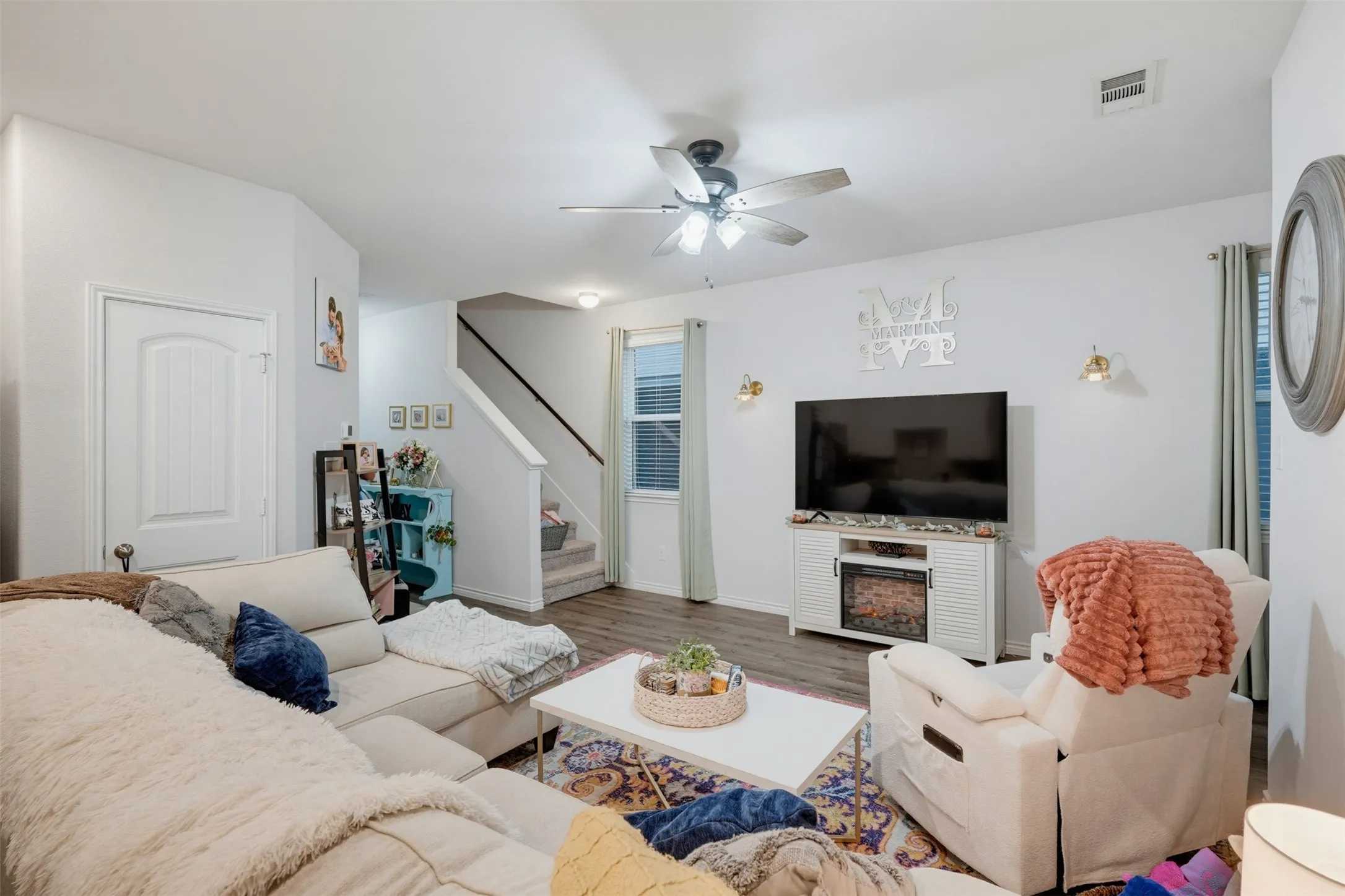 Living area with wood-finish flooring, a ceiling fan with integrated lighting, and a window with blinds
