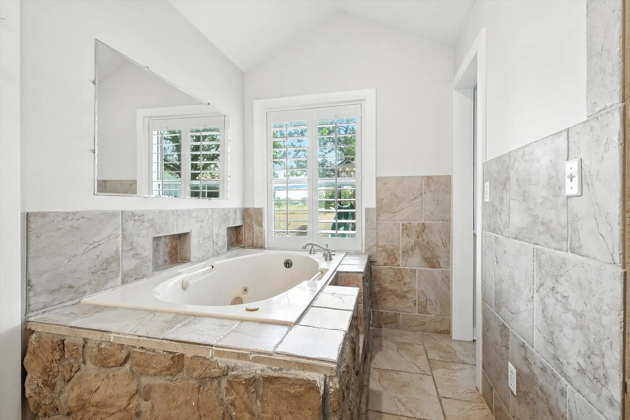 Built-in jetted tub surrounded by stone and tile, featuring a mounted mirror, vaulted ceiling, and plantation-shuttered windows