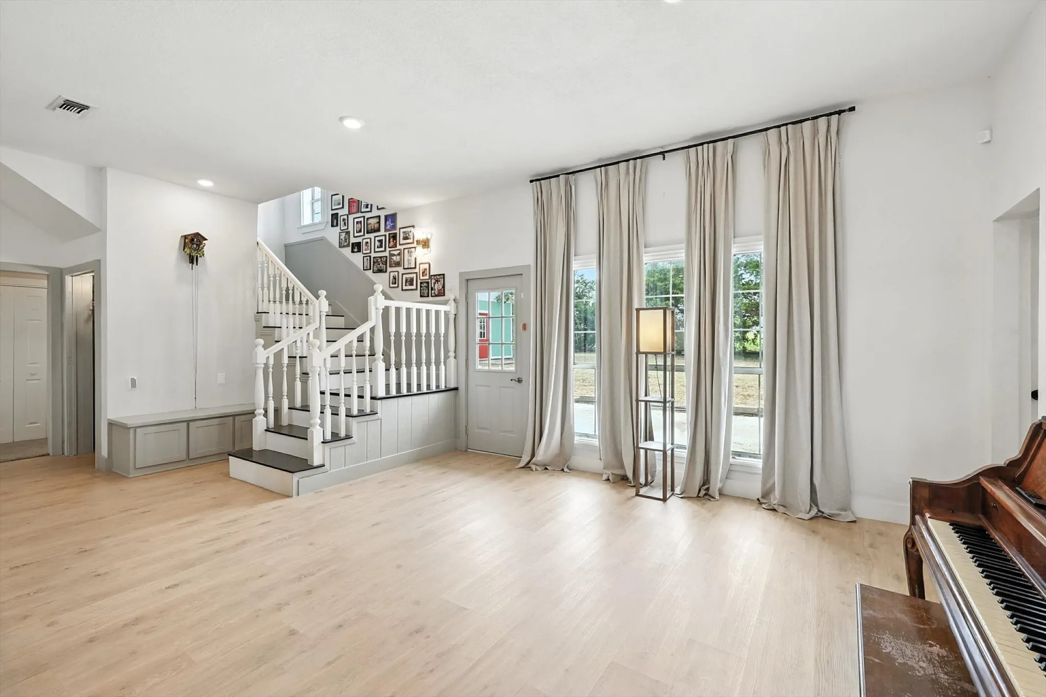 Light wood-finish flooring, staircase with white newel posts and handrails, three tall windows, recessed lighting, and built-in storage bench