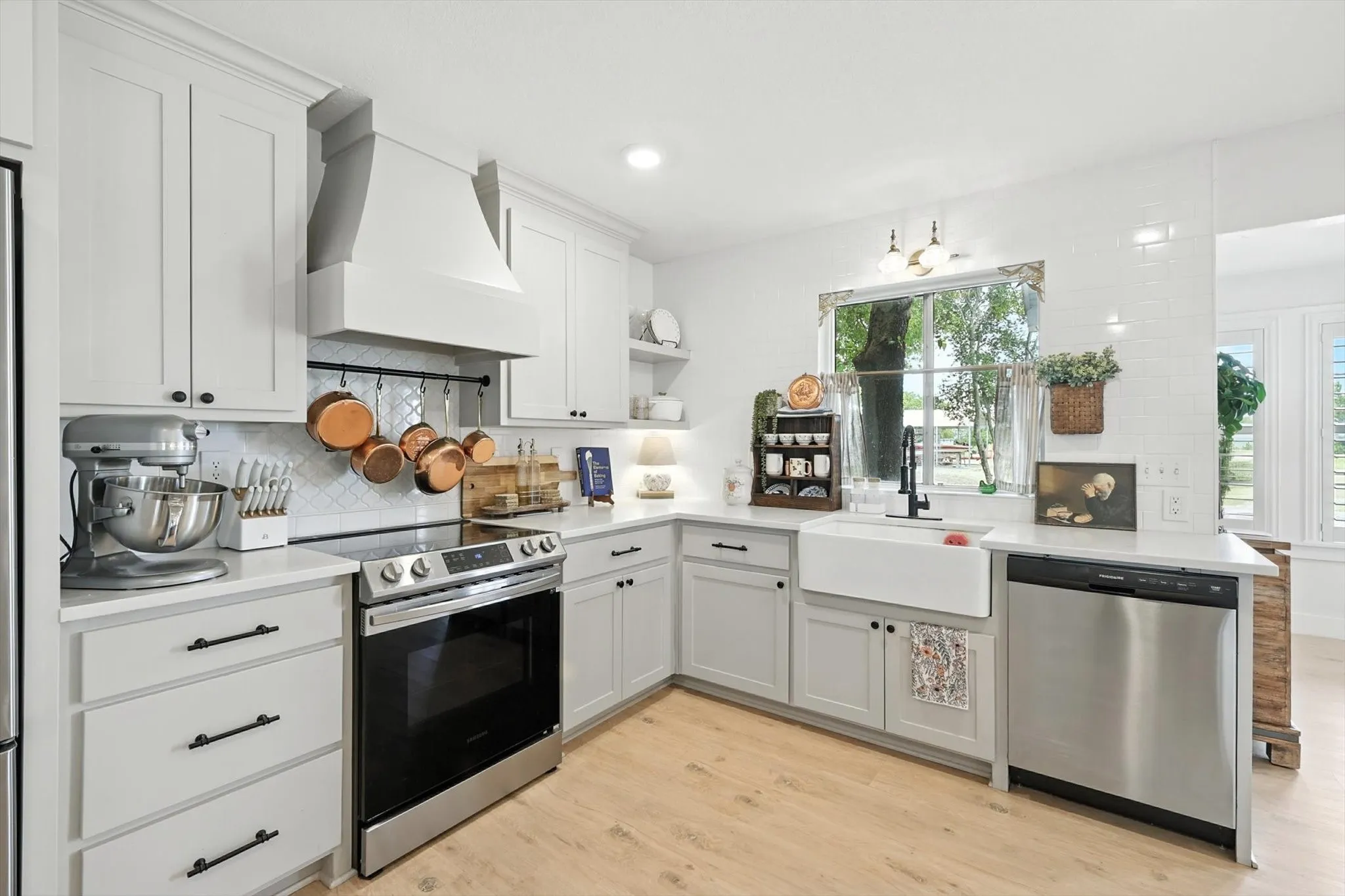 White shaker-style cabinetry with matte black hardware, stainless steel appliances, a farmhouse sink, and light wood-finish flooring