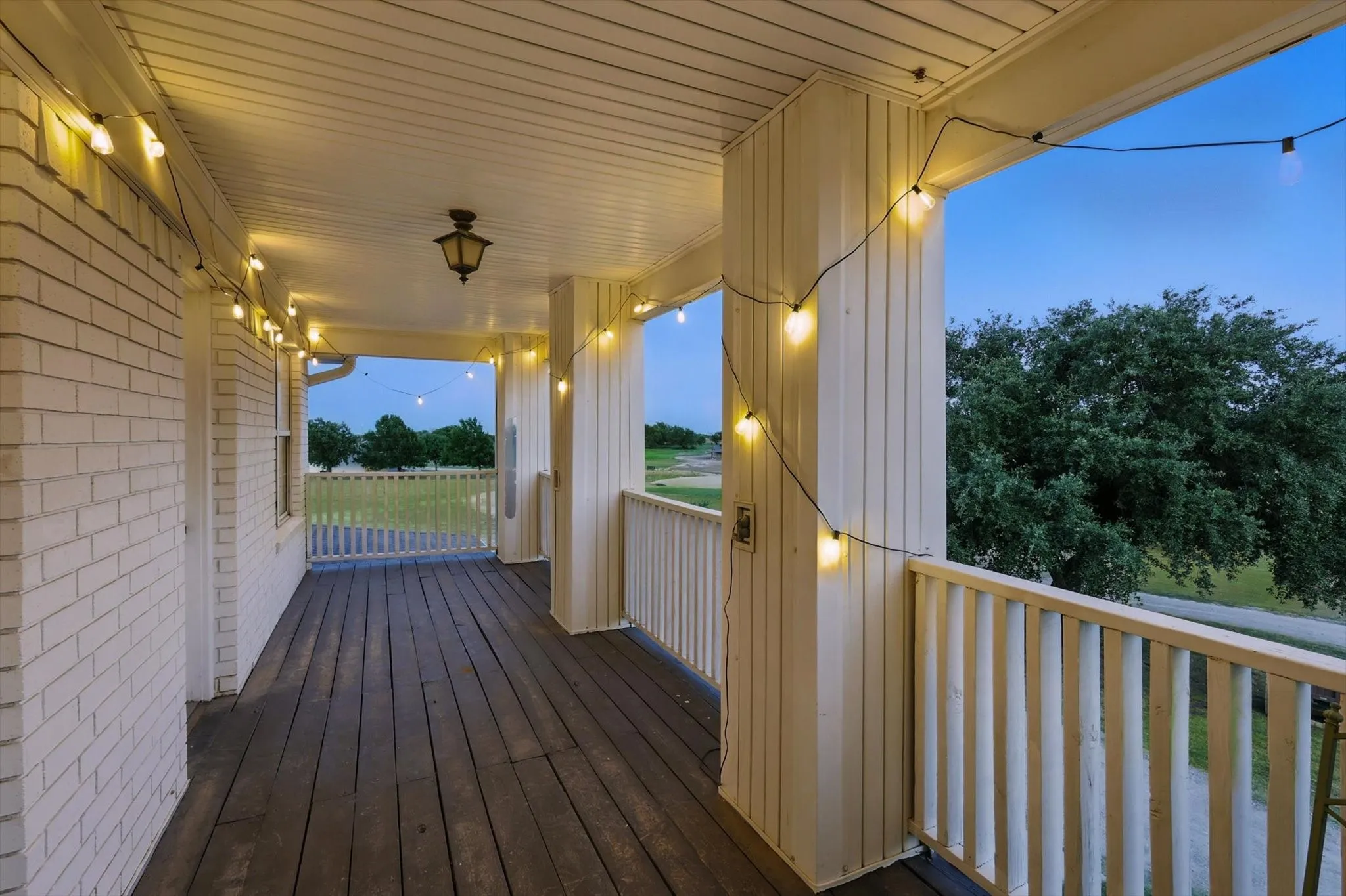 Covered porch featuring white brick exterior, wood-finish flooring, white railing, and a mounted ceiling light fixture
