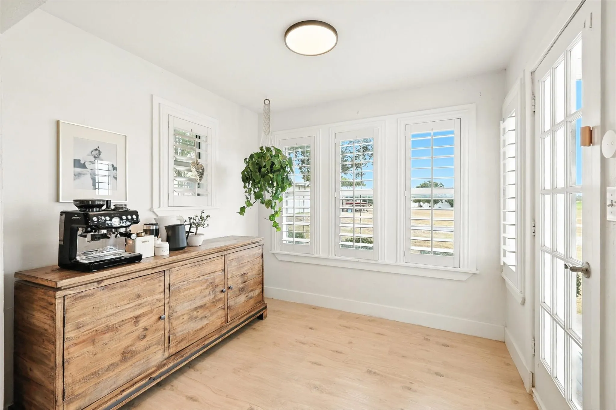 Light wood-finish flooring, white walls, multiple windows with plantation shutters, ceiling-mounted flush light fixture, and a French-style door with glass panels