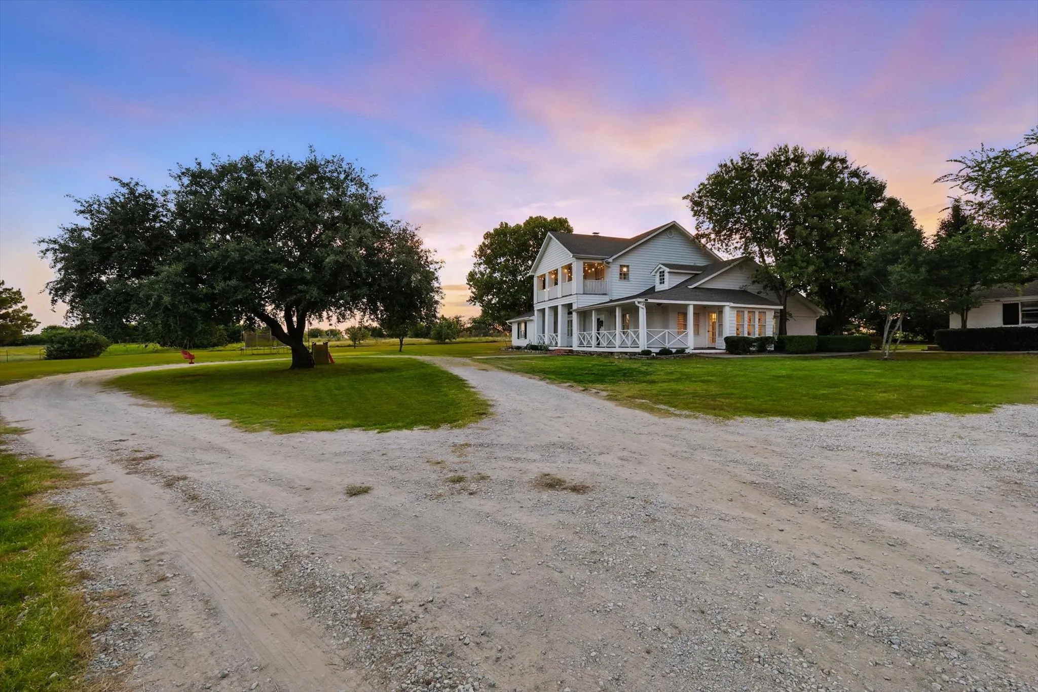 Expansive gravel drive leading to a classic two-story residence with a wraparound porch and white exterior siding