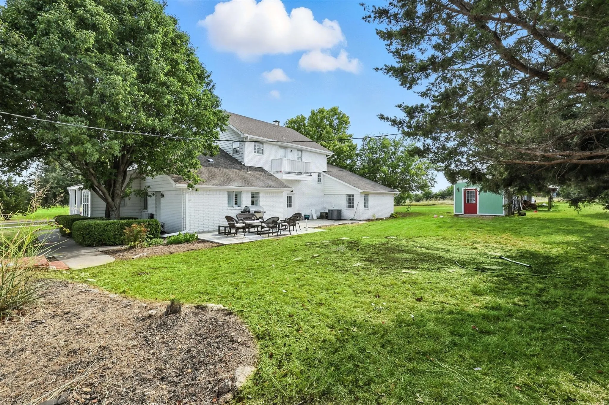 White brick exterior, upper-level balcony, expansive green lawn, mature trees, concrete patio