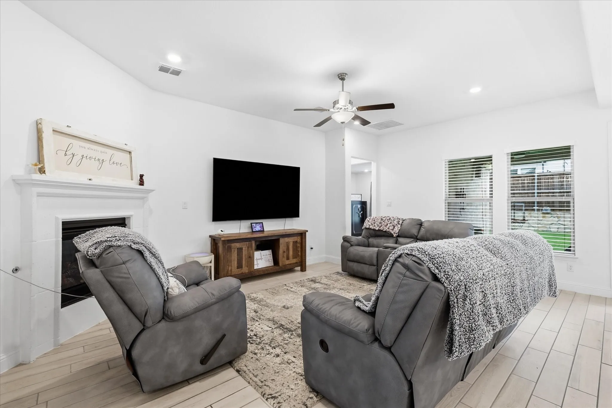 White walls with recessed lighting, a fireplace with a white mantel, a ceiling fan, multiple windows with blinds, and wood-finish flooring
