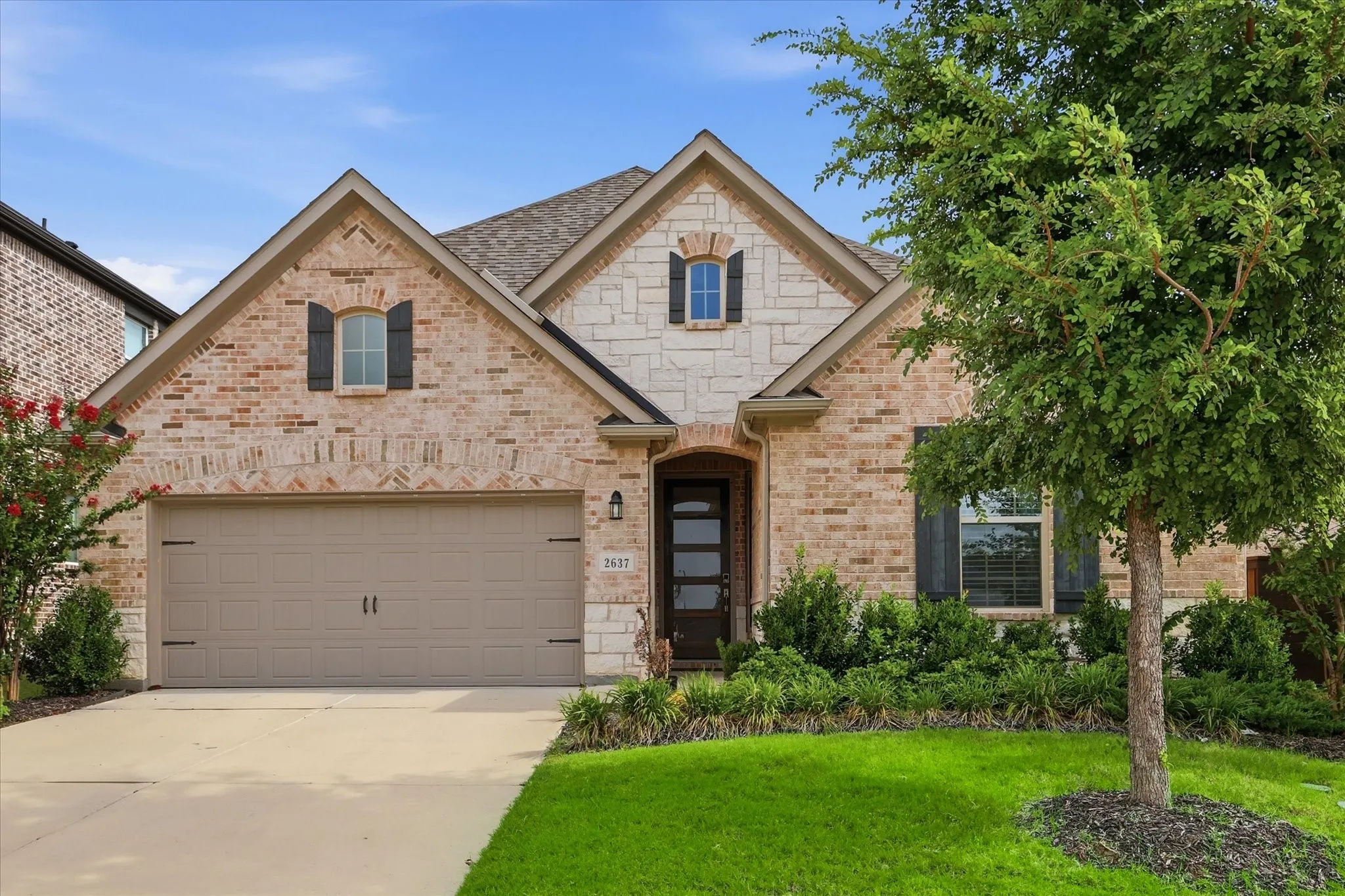 Brick and stone facade with dark shutters, an integrated two-car garage, and a mature tree