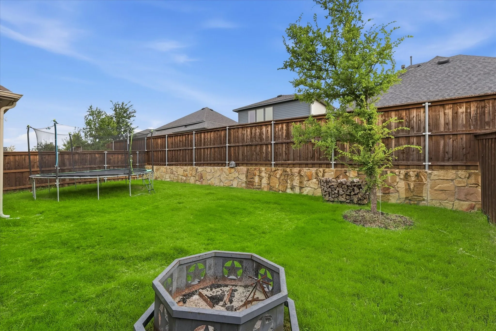 Expansive backyard featuring a manicured lawn, wood privacy fencing, stone retaining wall, and a fire pit