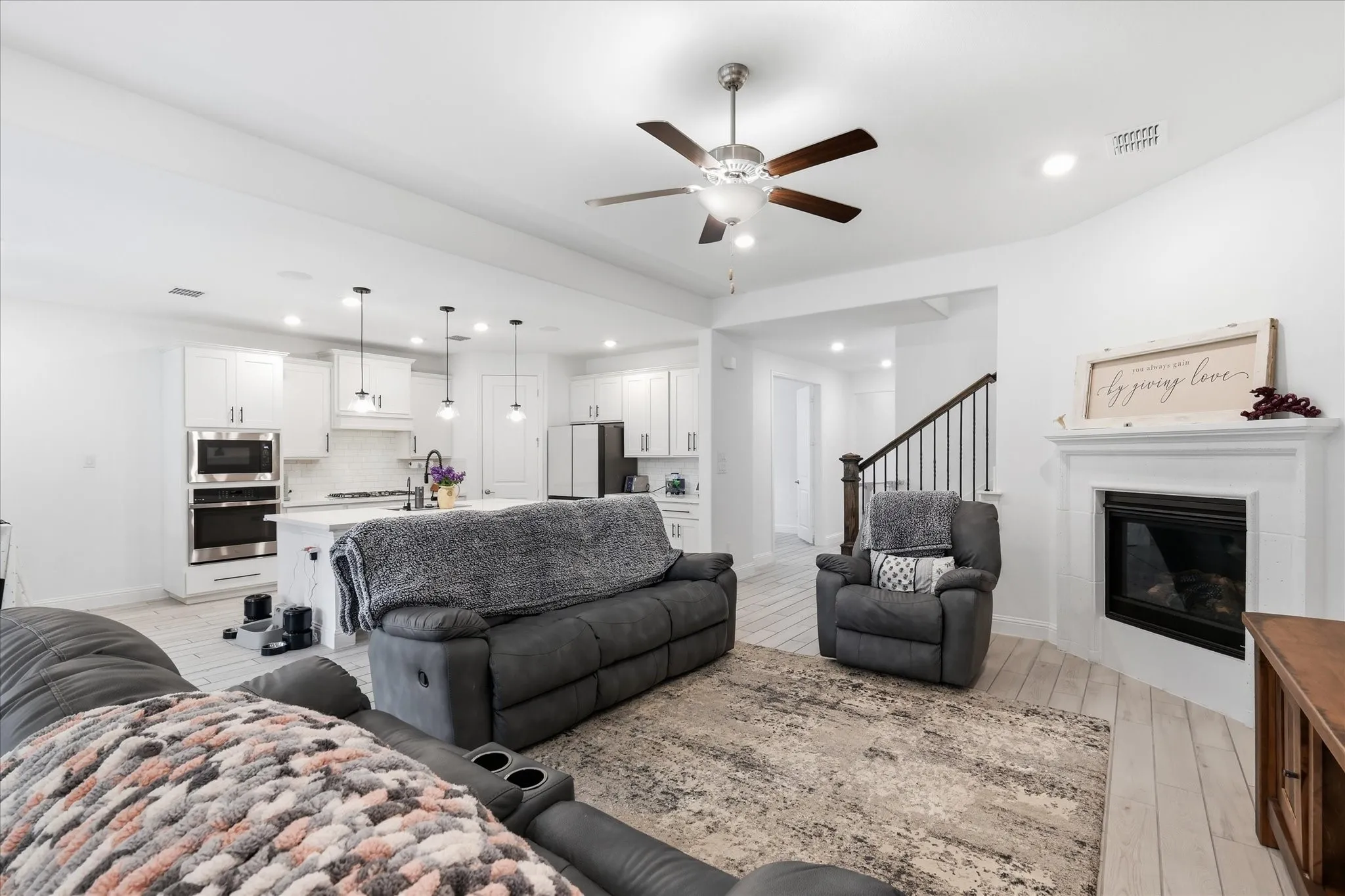 Open-concept living area featuring light wood-finish flooring, a contemporary ceiling fan, and a fireplace with a white mantel