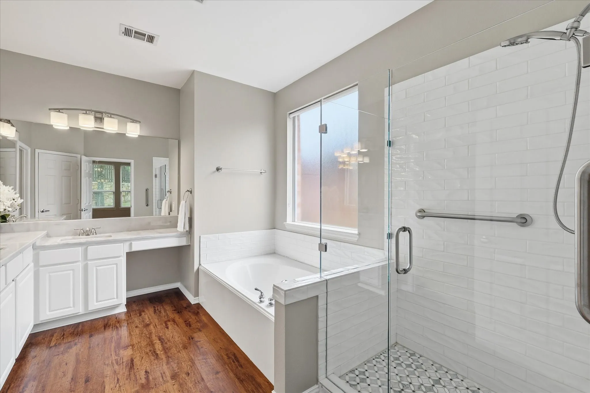 Wood-finish flooring, white vanity with dual sinks, built-in soaking tub, glass-enclosed shower with subway tile, and frosted window