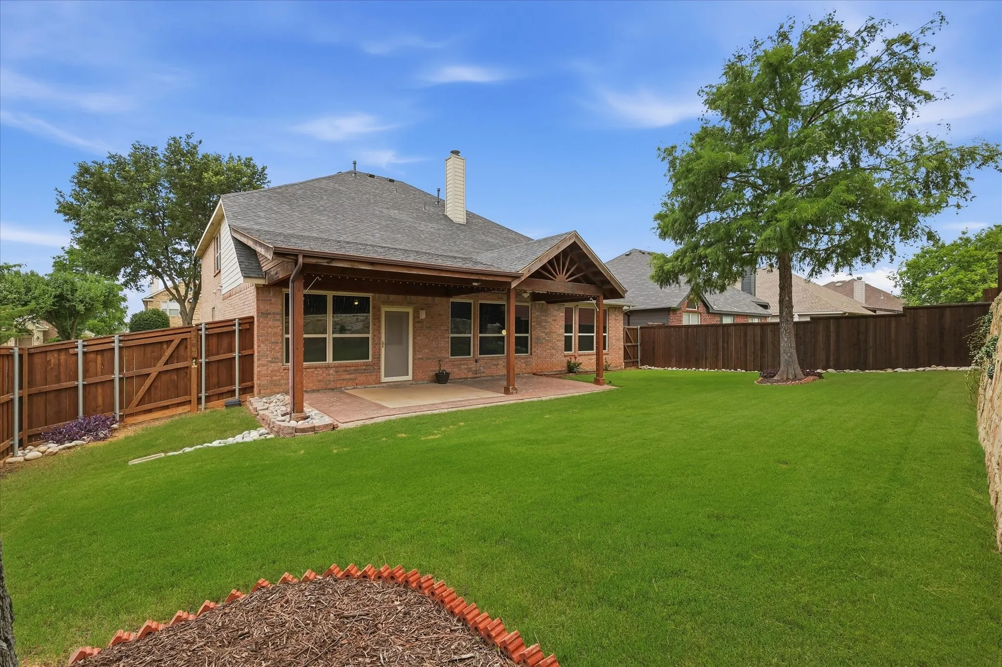 Expansive backyard featuring a manicured lawn, full privacy fence, mature trees, and a brick patio with an attached covered outdoor living area