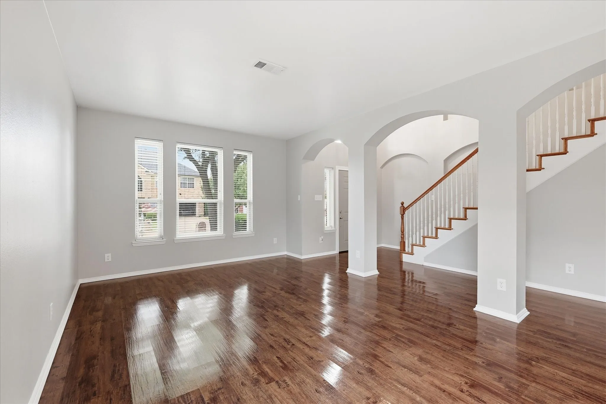 Expansive room featuring rich wood-finish flooring, multiple windows with blinds, a grand staircase with wood handrails, and arched doorways