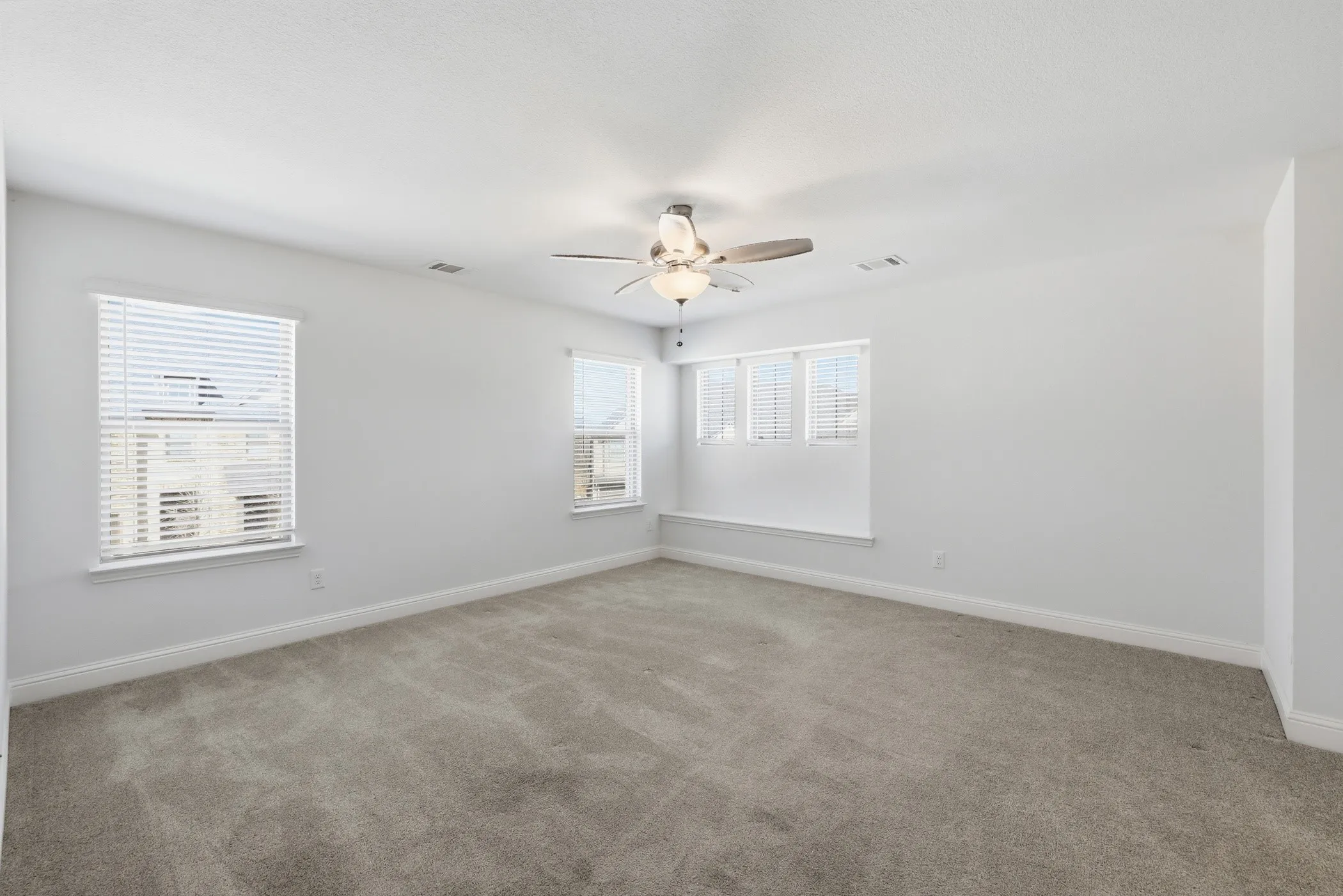 Expansive room featuring light gray carpeting, multiple windows with white blinds, a built-in window bench, and a ceiling fan with integrated lighting