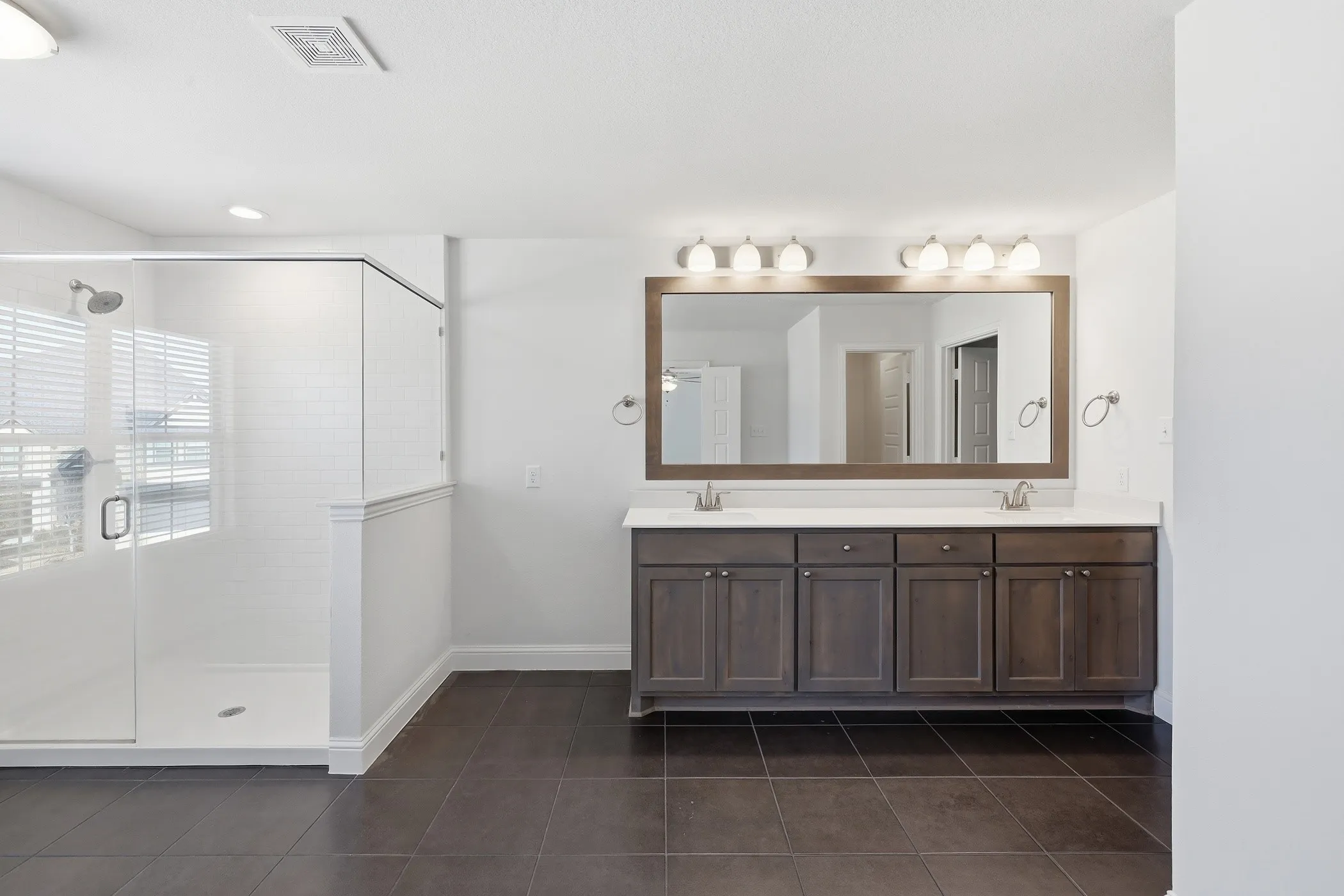 Glass-enclosed shower with white subway tile, dual vanity with wood-finish cabinetry, rectangular mirror, and dark tile flooring