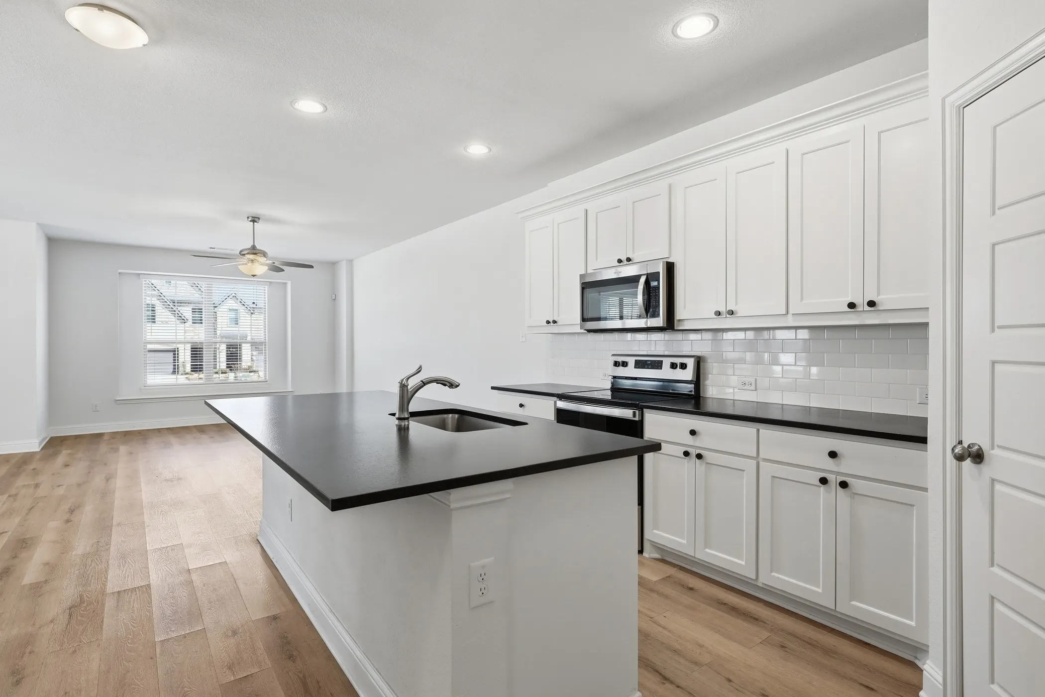 Kitchen island with an undermount sink, white shaker-style cabinetry, dark countertops, stainless steel appliances, and wood-finish flooring