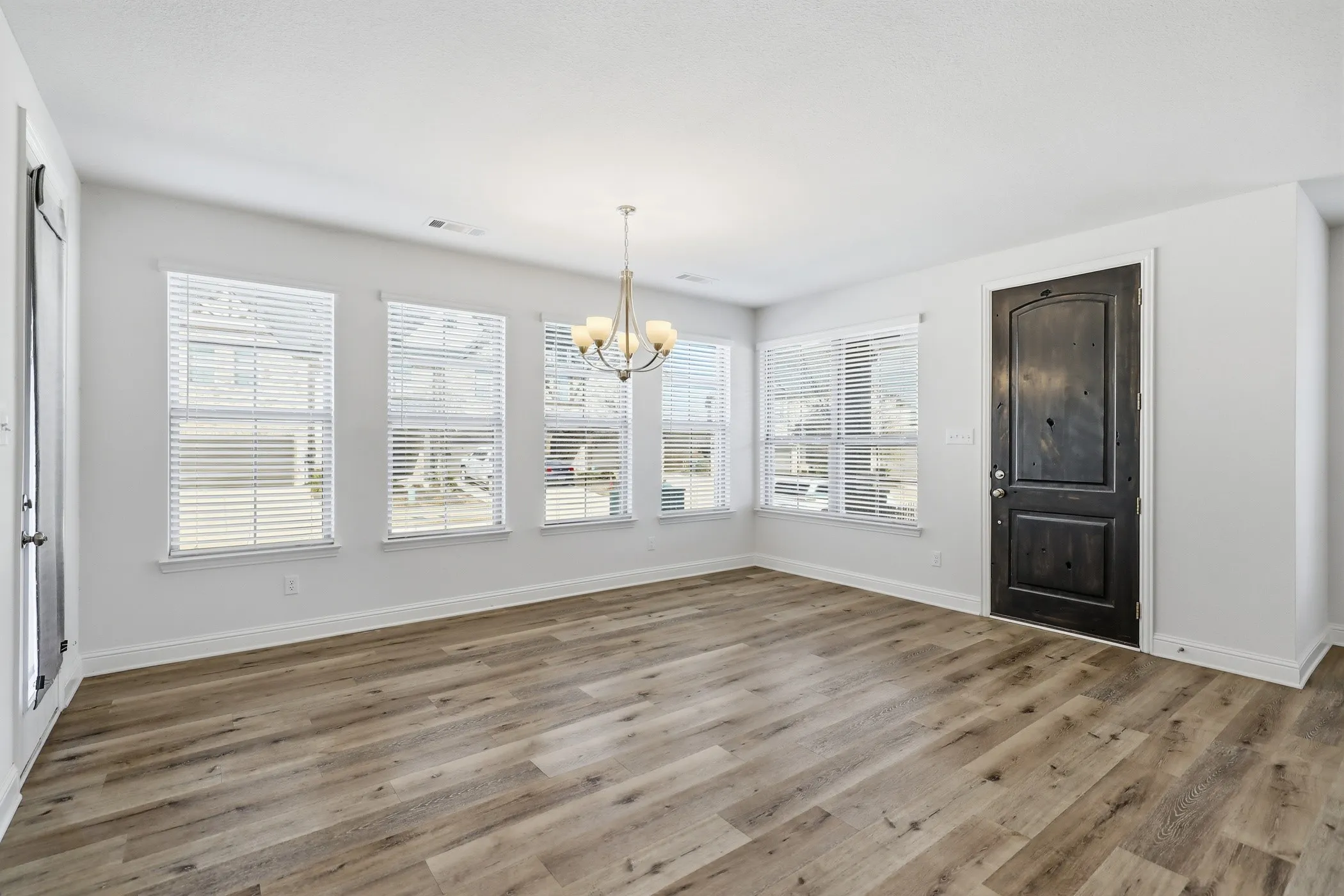 Wood-finish flooring, multiple windows with blinds, a dark wood-paneled door, and a multi-bulb chandelier