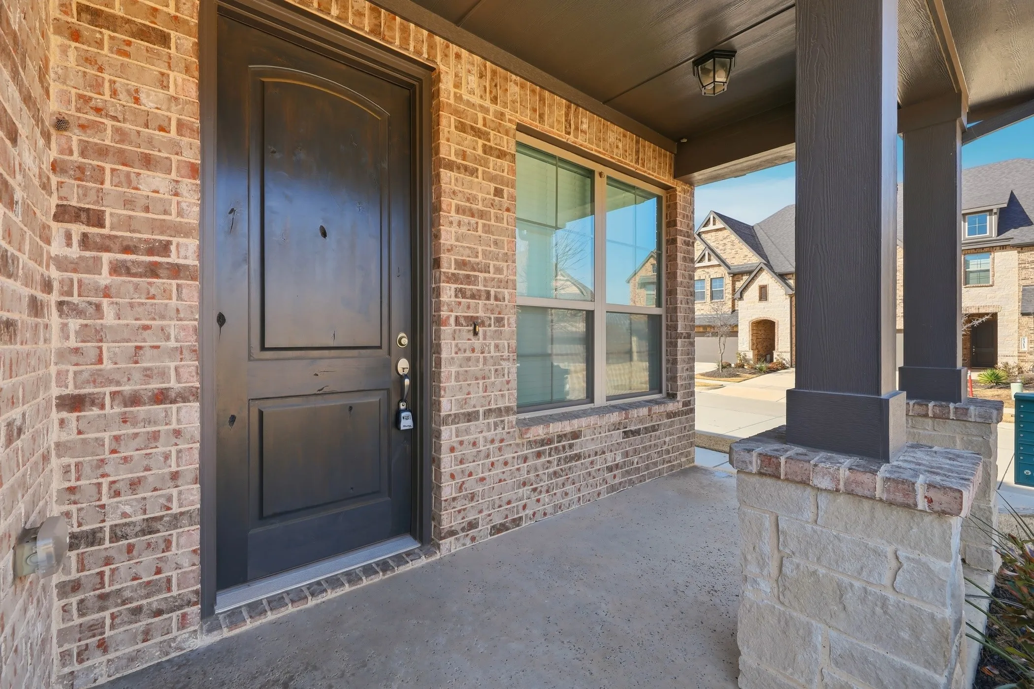 Covered front porch featuring a dark wood-finish entry door, brick exterior, stone-clad columns, and an integrated ceiling light​​‌​​​​‌​​‌‌​‌‌​​​‌‌​‌​‌​‌​​​‌​​ fixture