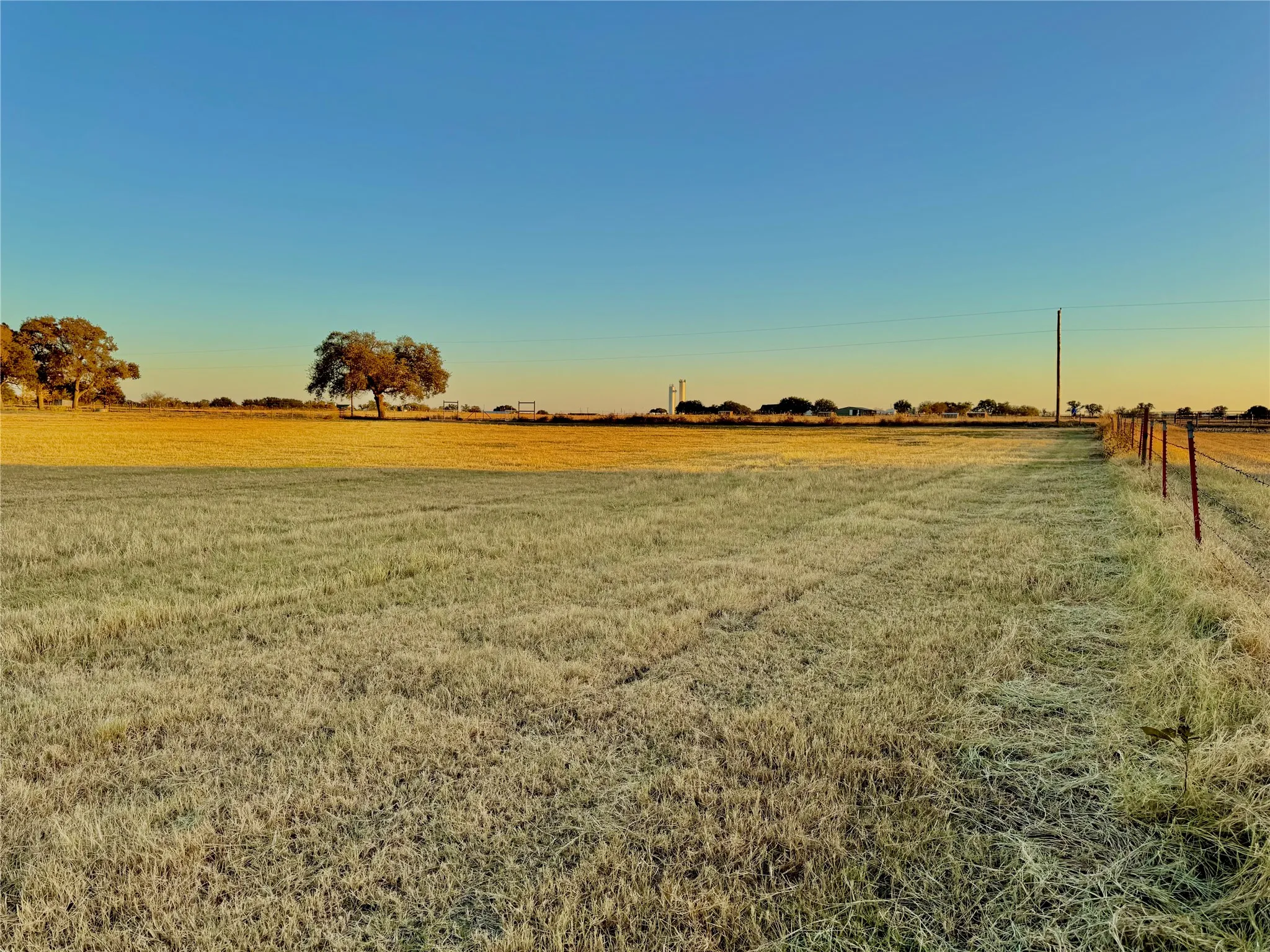 Expansive acreage with a wire fence line, mature trees, and clear sky