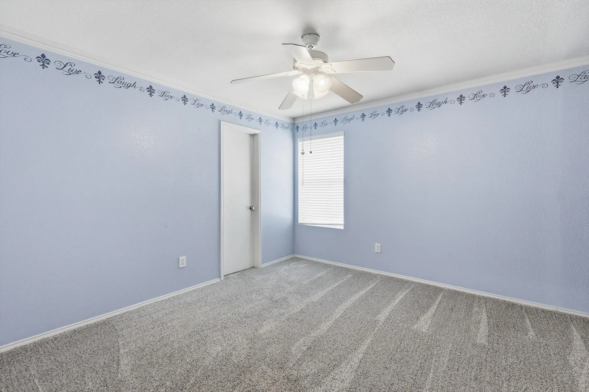 Bright secondary bedroom featuring soft periwinkle walls, decorative border trim, neutral carpet flooring, ceiling fan with integrated lighting, and abundant natural light from a large window with blinds