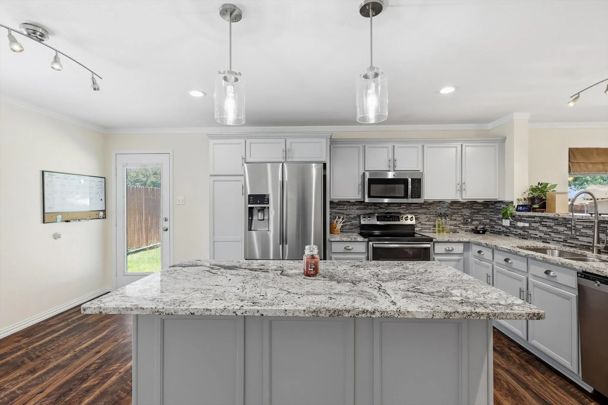 Chef's kitchen featuring custom gray cabinetry, granite countertops, a large waterfall-edge island with seating, stainless steel appliances including a French-door refrigerator and gas range, mosaic tile backsplash, pendant lighting, and hardwood flooring throughout the open layout with direct access to the backyard