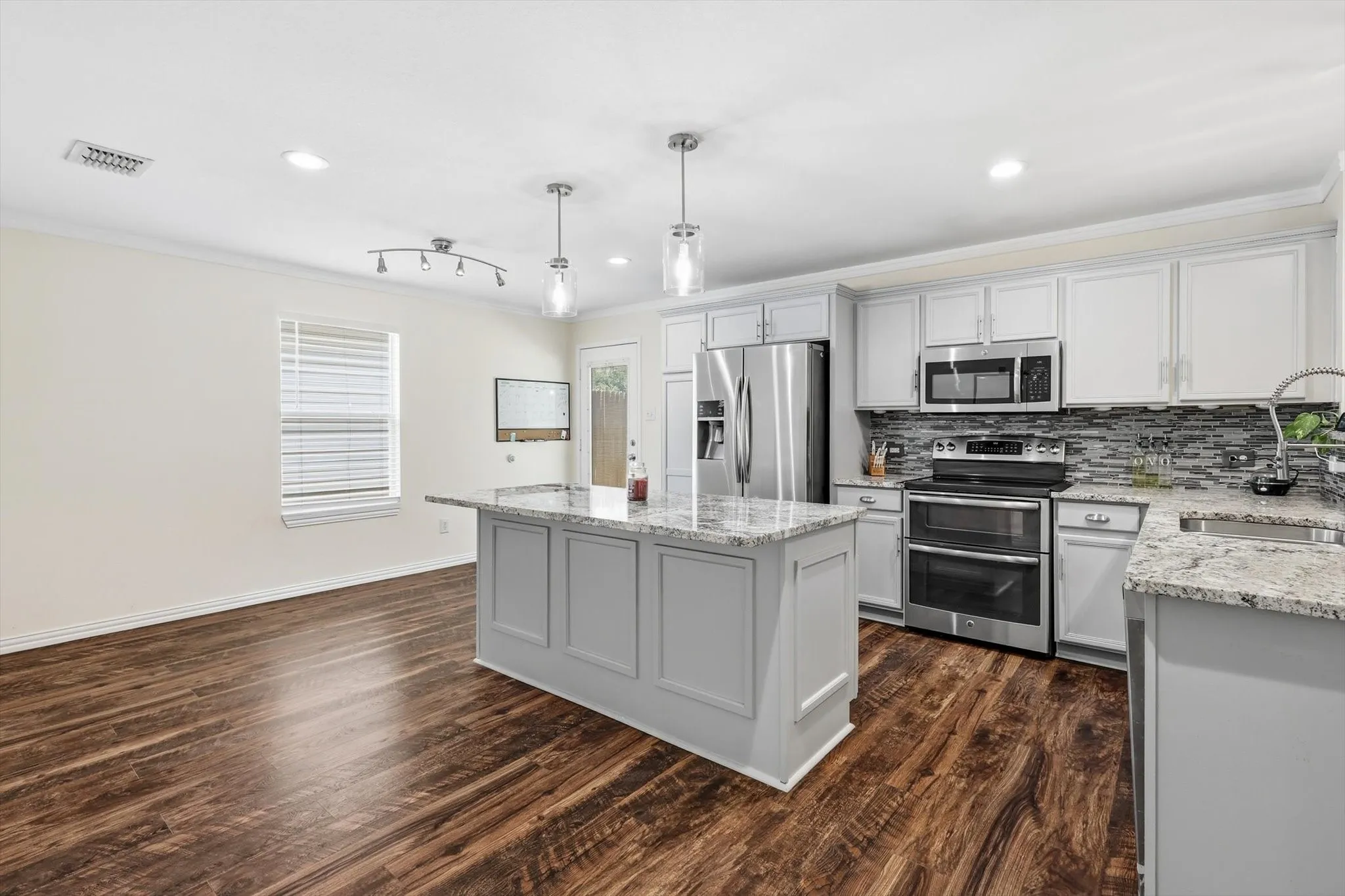 Modern kitchen featuring white cabinetry with a large center island, stainless steel appliances including a double wall oven and microwave, granite countertops, and a tile backsplash
