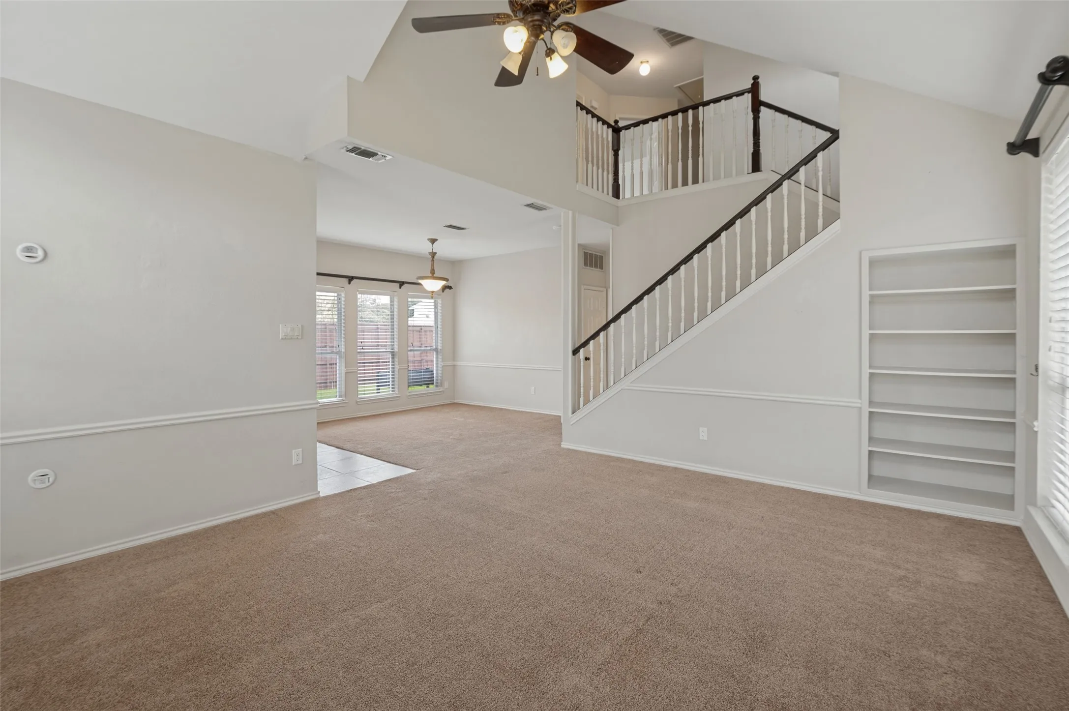 Vaulted ceiling, ceiling fan, dual-tone staircase with white balusters, built-in shelving, and light-colored carpeting