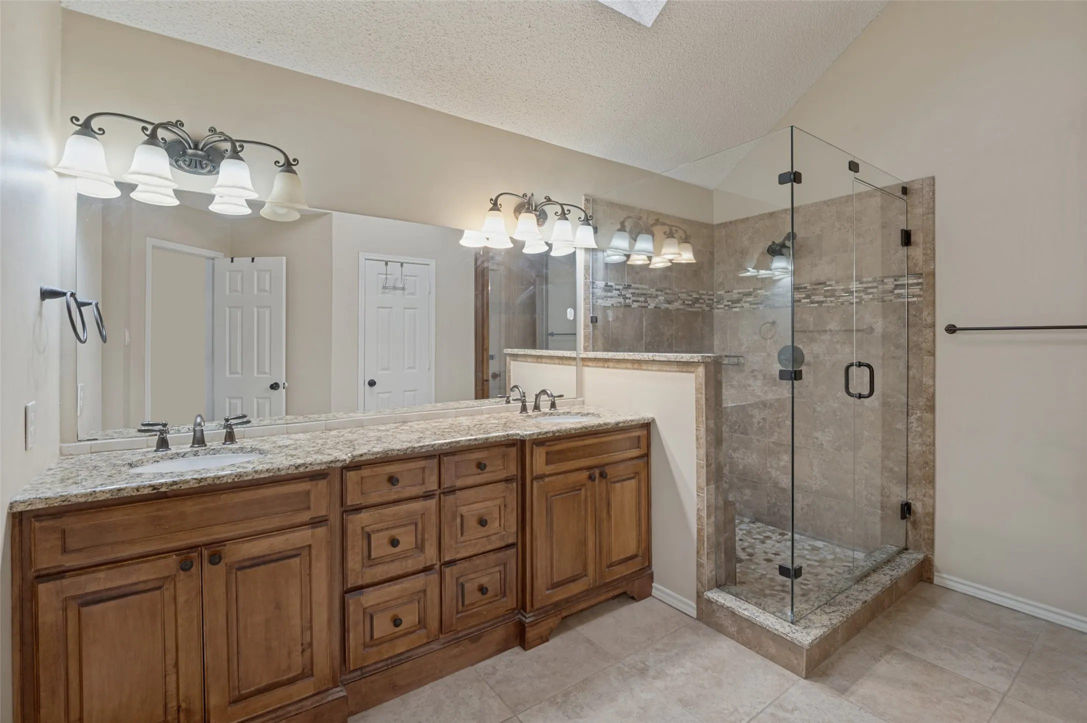 Dual sink vanity with wood-finish cabinetry, a large frameless mirror, and overhead sconce lighting